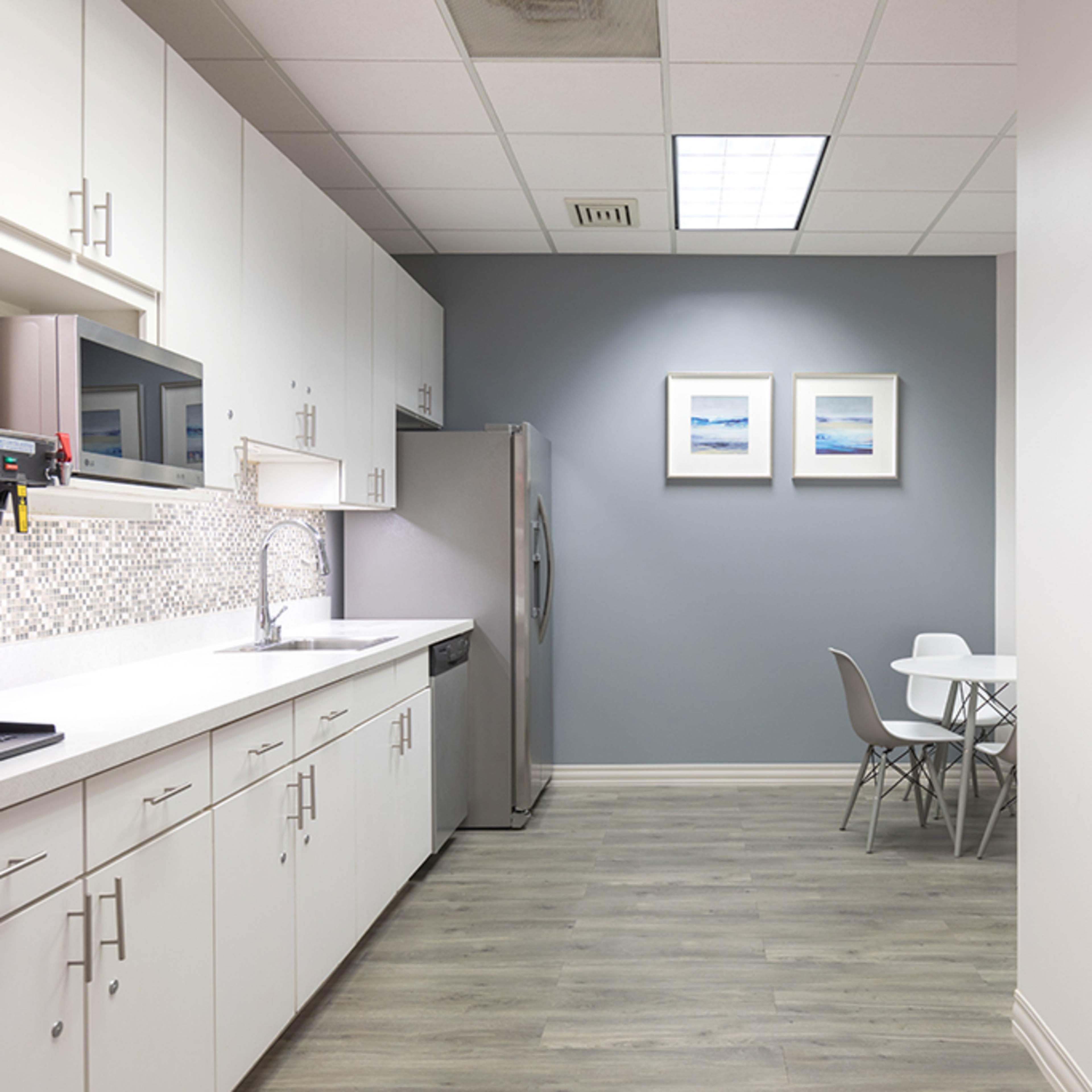 A modern kitchen area with white cabinetry, stainless steel appliances, a small dining table, and light grey walls adorned with two framed pictures.
