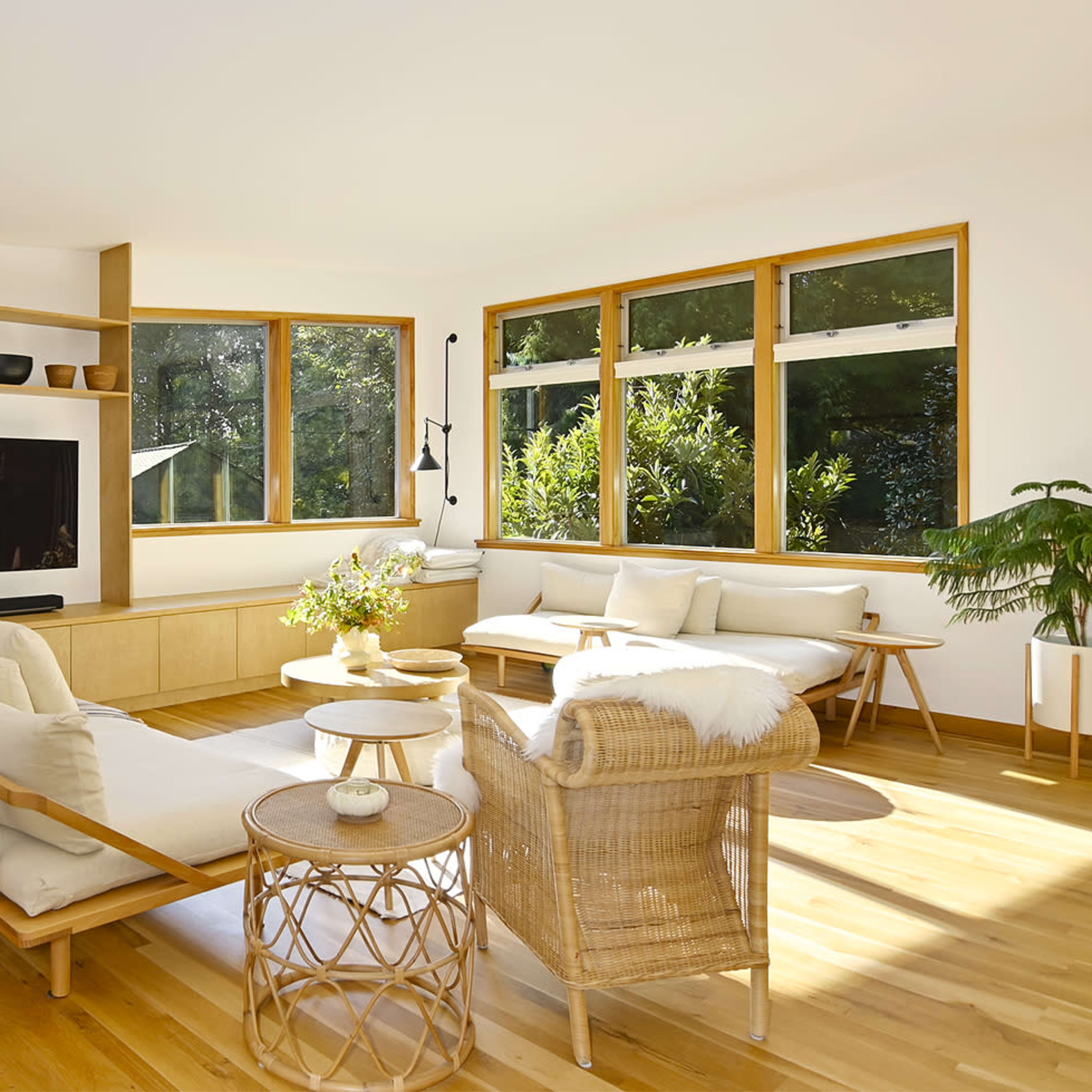 The room features a light-filled living area with wooden flooring, a minimalist shelf display, a television, and comfortable seating arrangements.