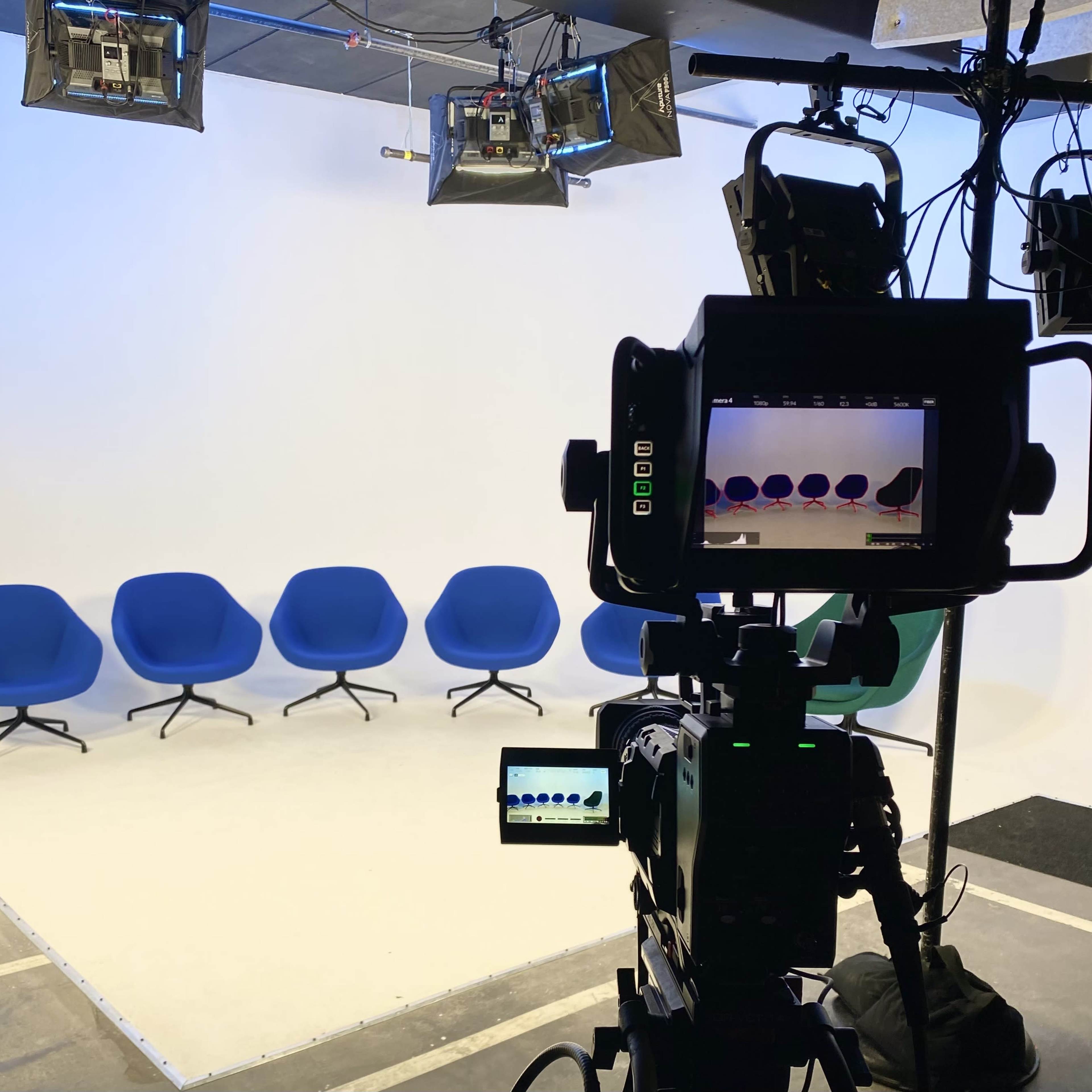 A camera setup faces a row of five blue chairs against a white backdrop in a studio.