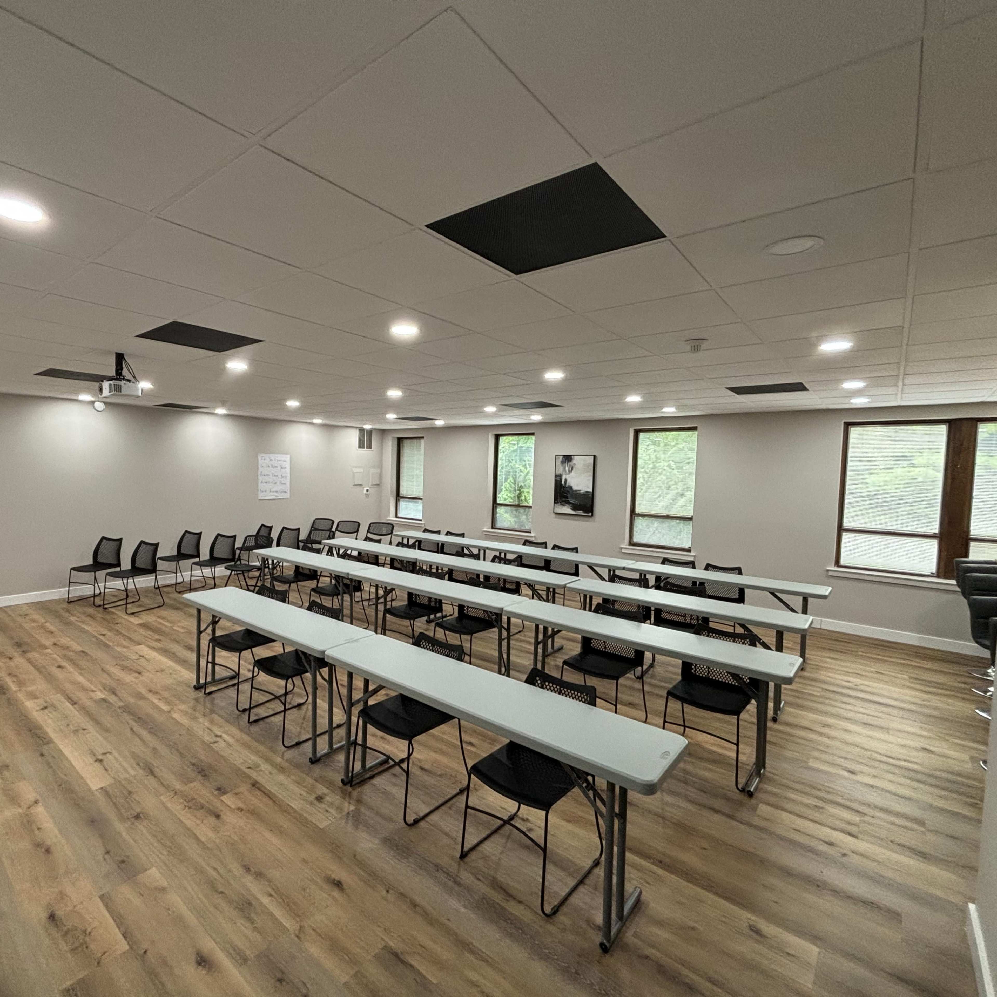 A modern conference room features rows of tables and chairs, large windows, and a wooden floor.