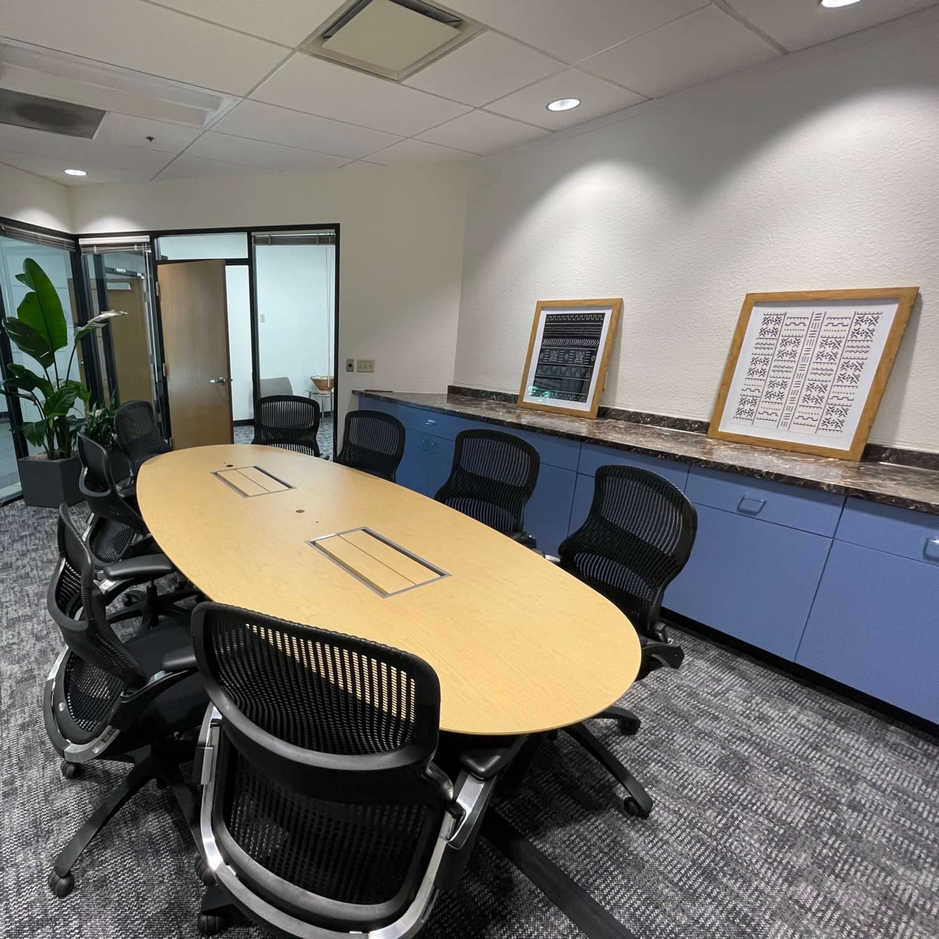 A conference room with an oval table surrounded by black chairs, adjacent to glass-walled office spaces.