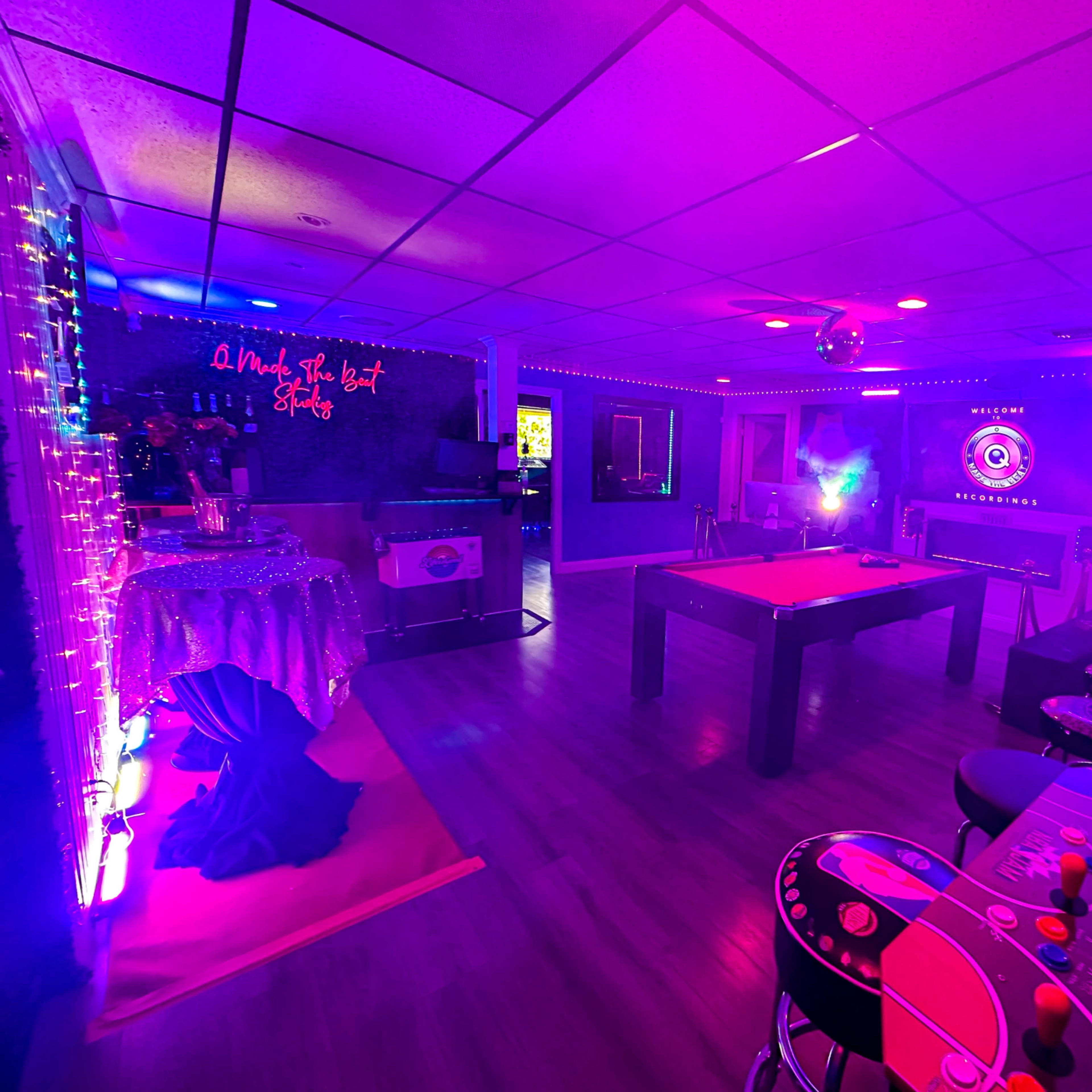 A dimly lit game room featuring a pool table, arcade machine, bar area, and various colorful lights.