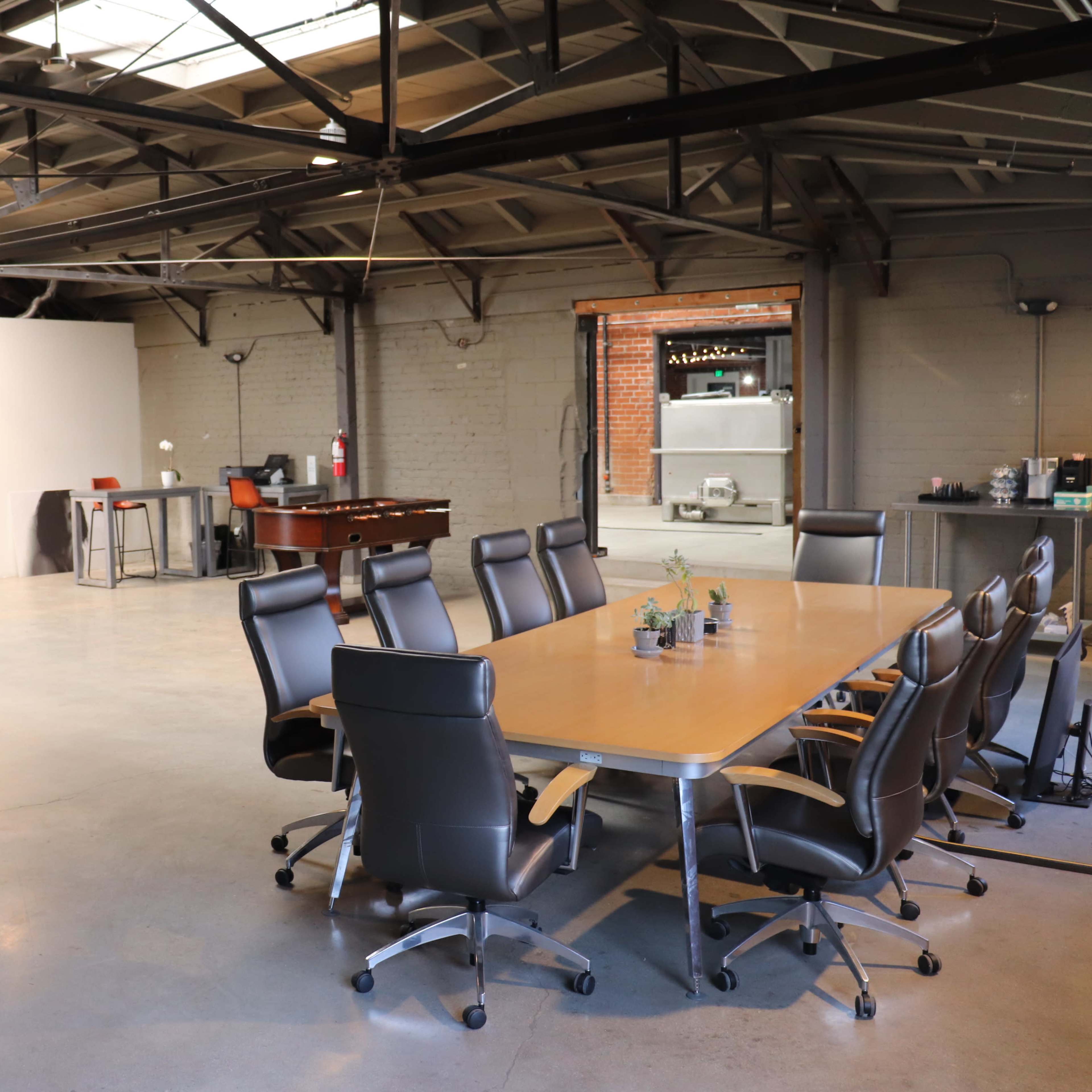 A spacious industrial-style office featuring a large conference table surrounded by black chairs, with workstations and a kitchenette visible in the background.
