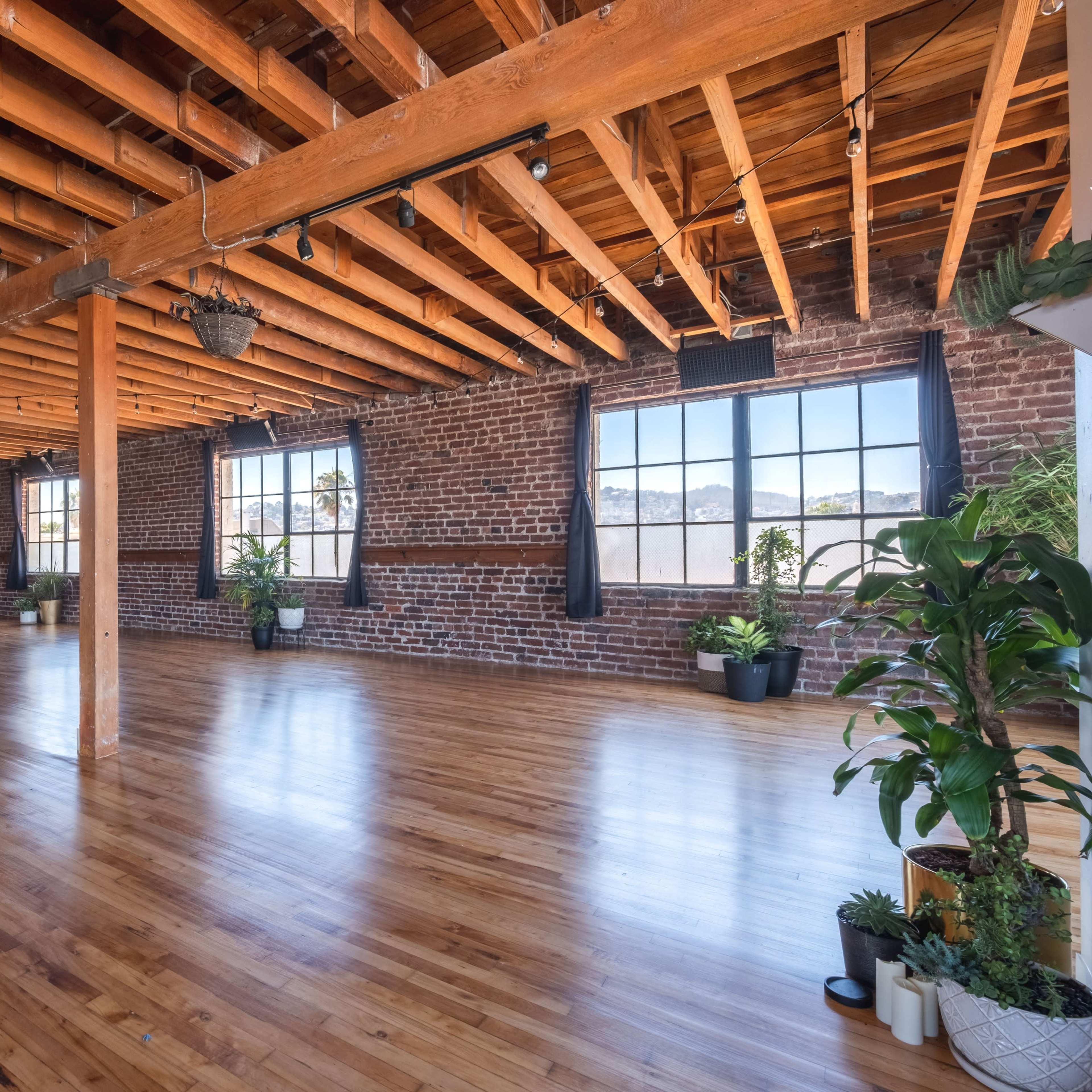A spacious indoor area featuring exposed wooden beams, brick walls, and large windows with plants positioned around the room.