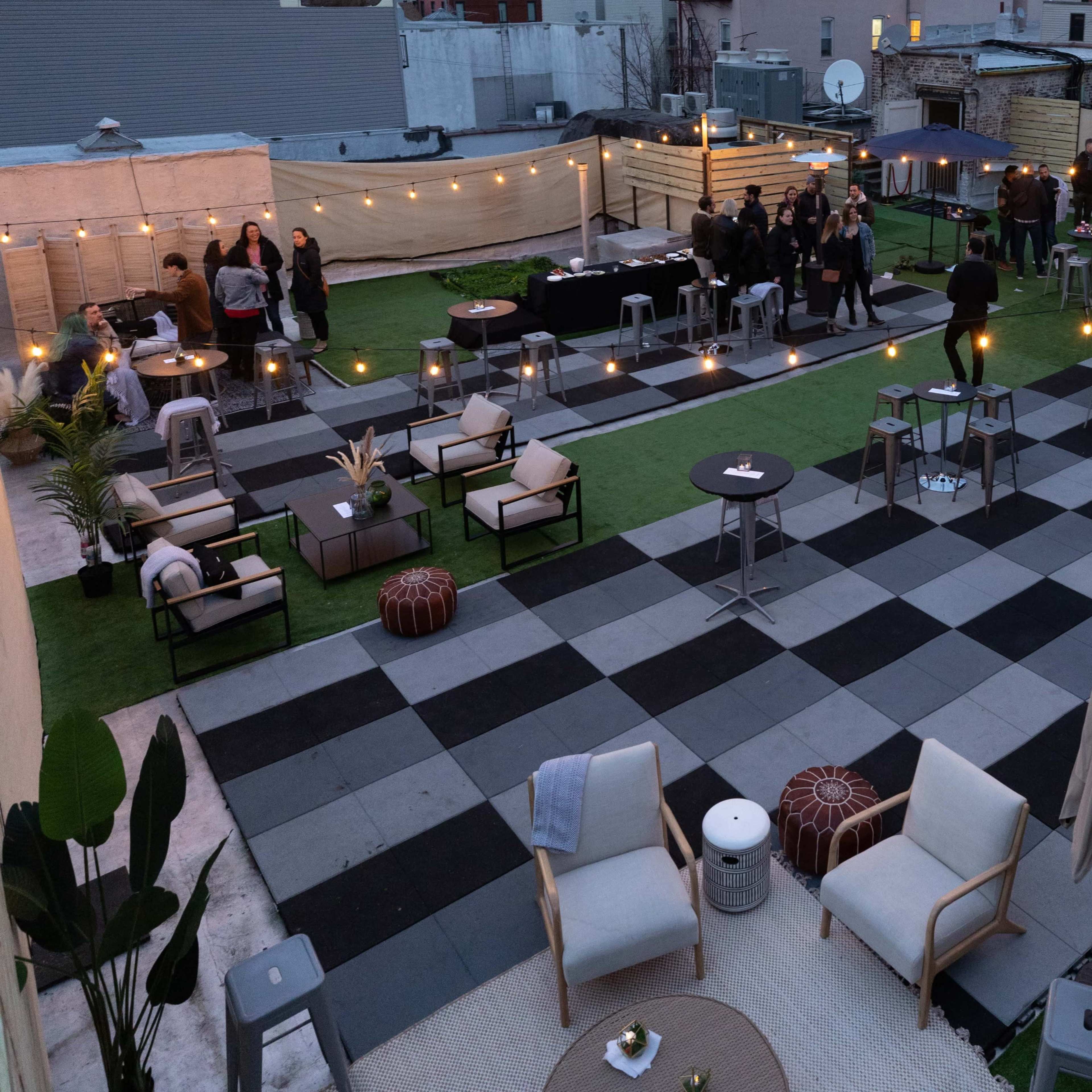 A rooftop space is set up for an event, featuring a checkerboard-patterned floor with scattered tables, chairs, and string lights overhead.
