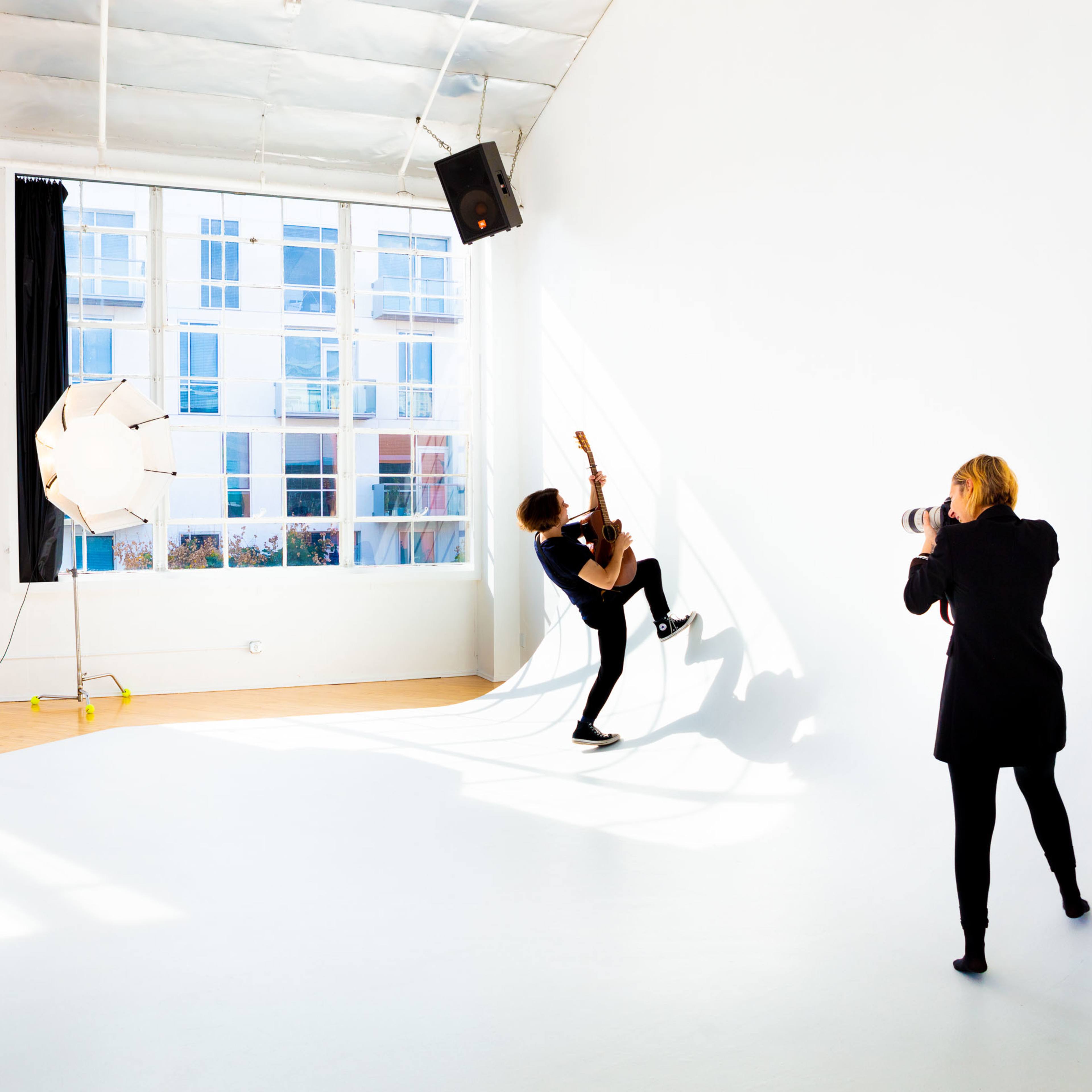 A person plays guitar against a white wall while another person photographs them in a brightly lit studio.