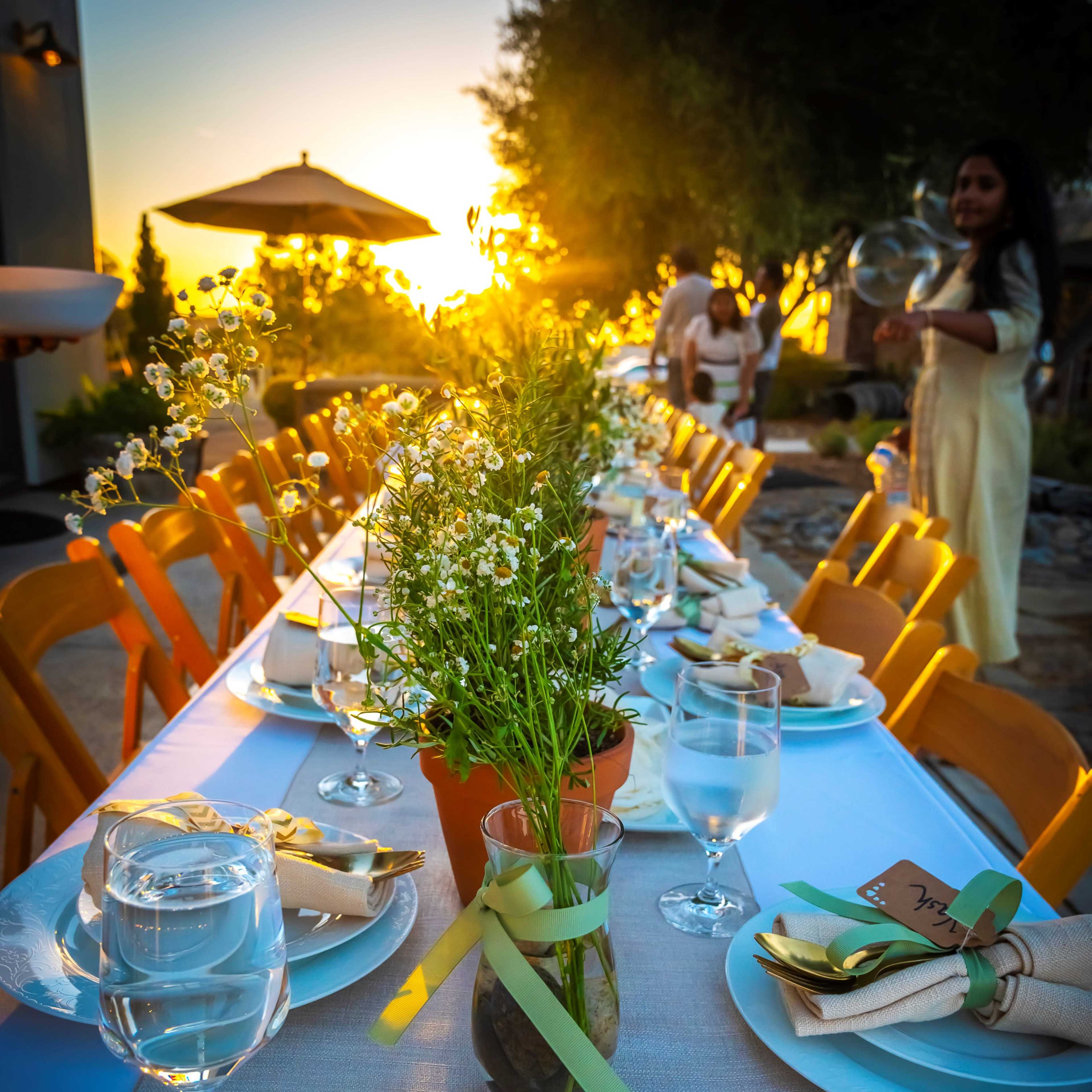 A long table set for an outdoor dinner features flower arrangements and glasses of water, with a sunset in the background.