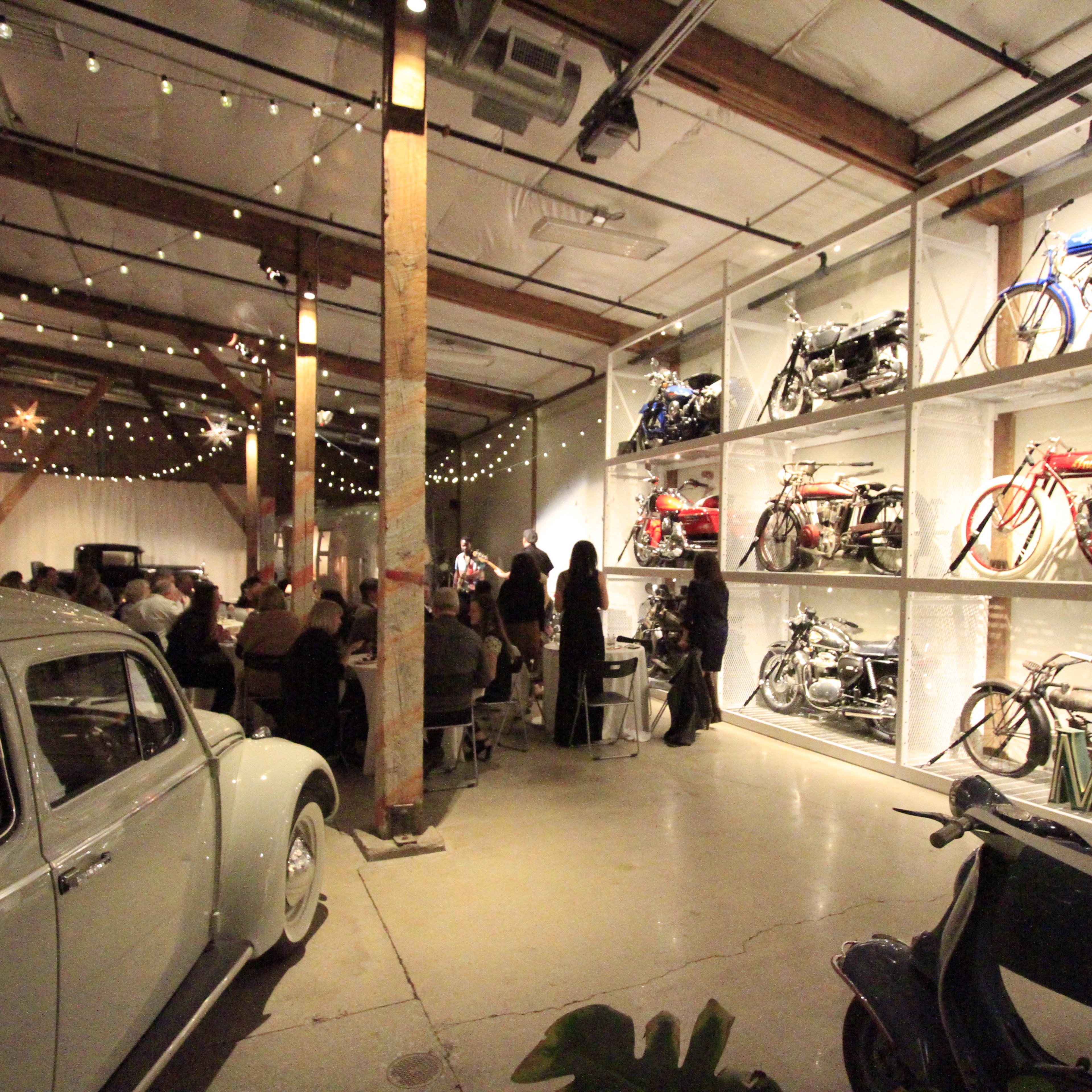 A large indoor space with decorative string lights, featuring vintage bicycles displayed on wall shelves alongside a classic car and tables where people are seated.
