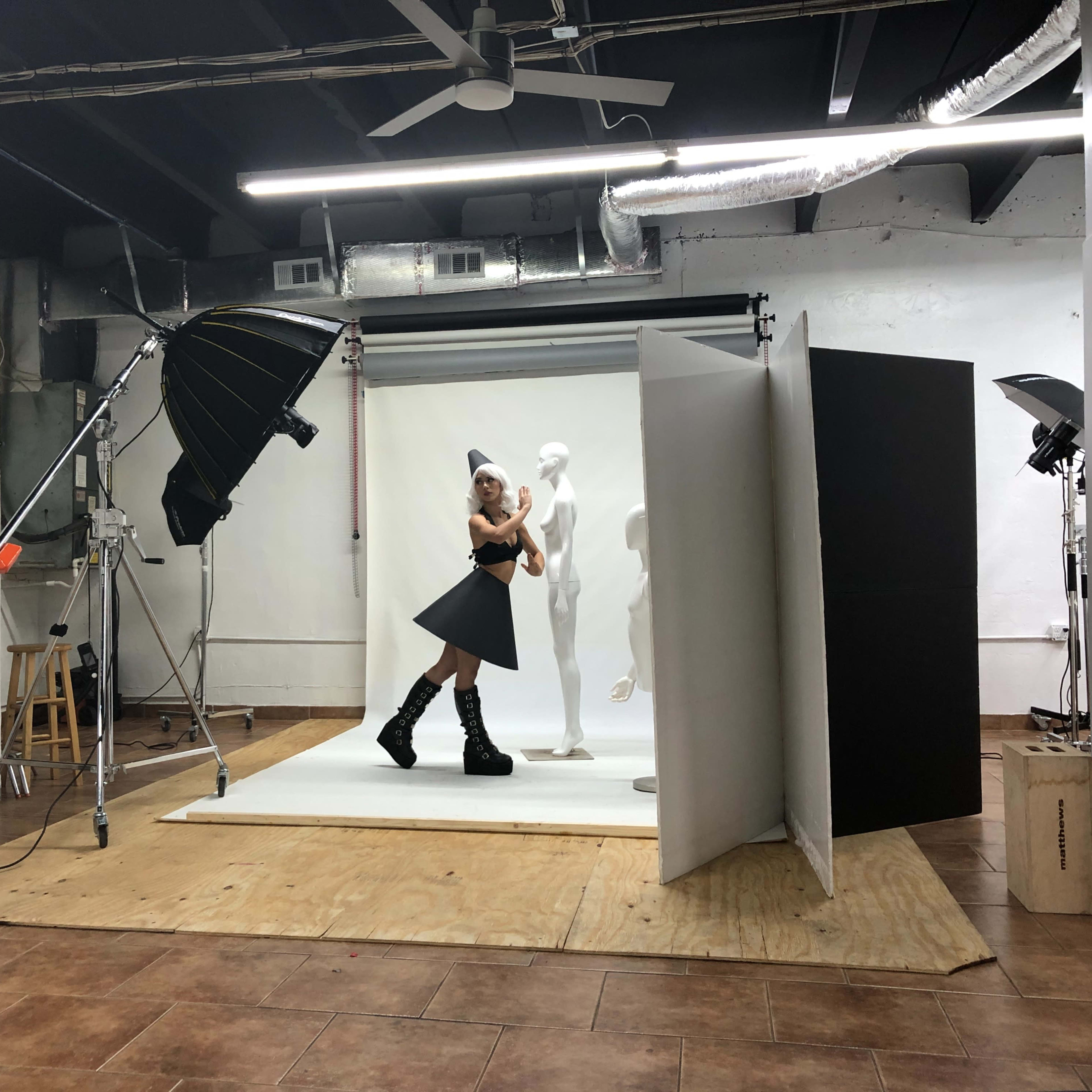 A model poses in front of a white backdrop in a photography studio, surrounded by lighting equipment and a mannequin.