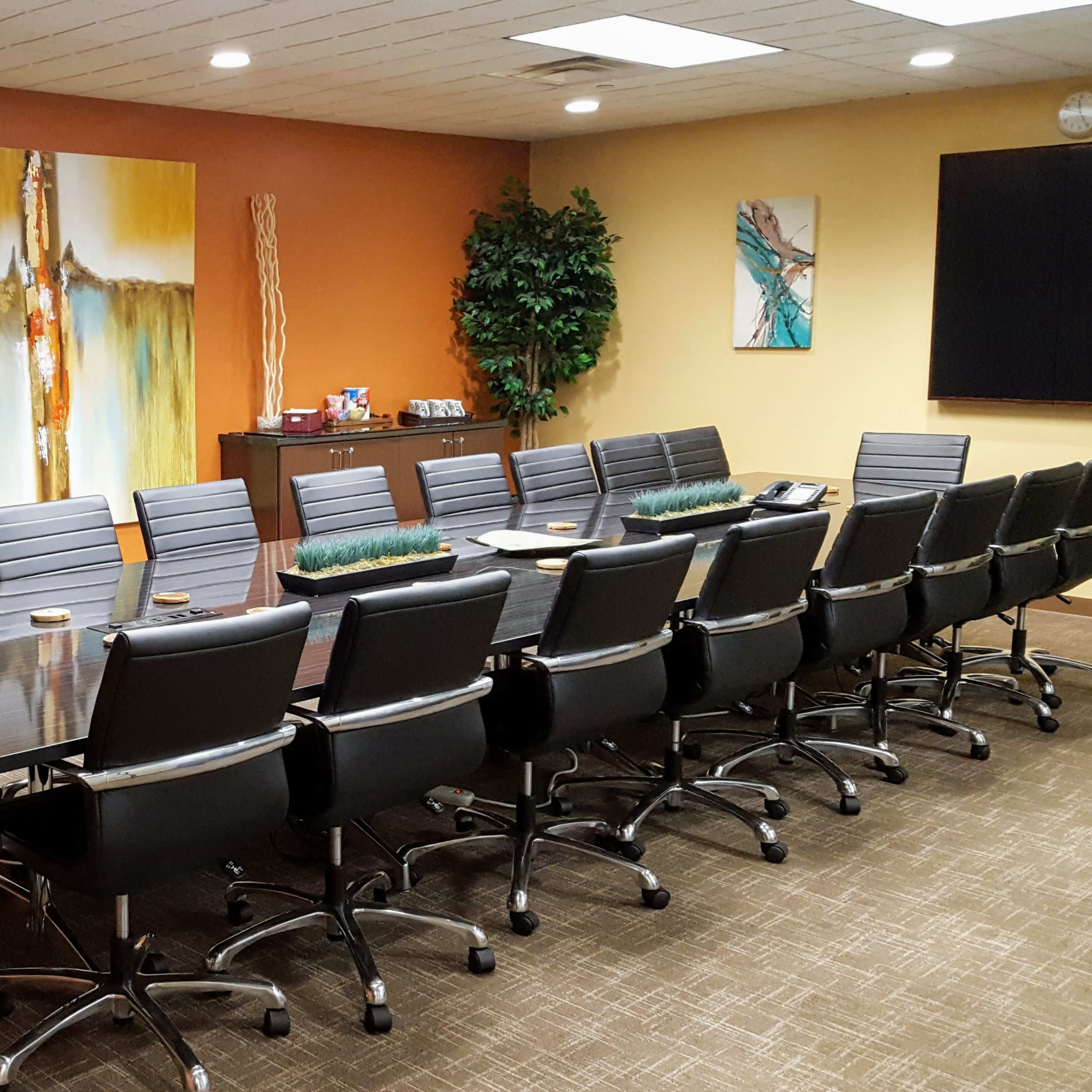 A modern conference room features a long black table surrounded by ergonomic chairs, with large artwork and a television displaying an aquarium scene on the wall.