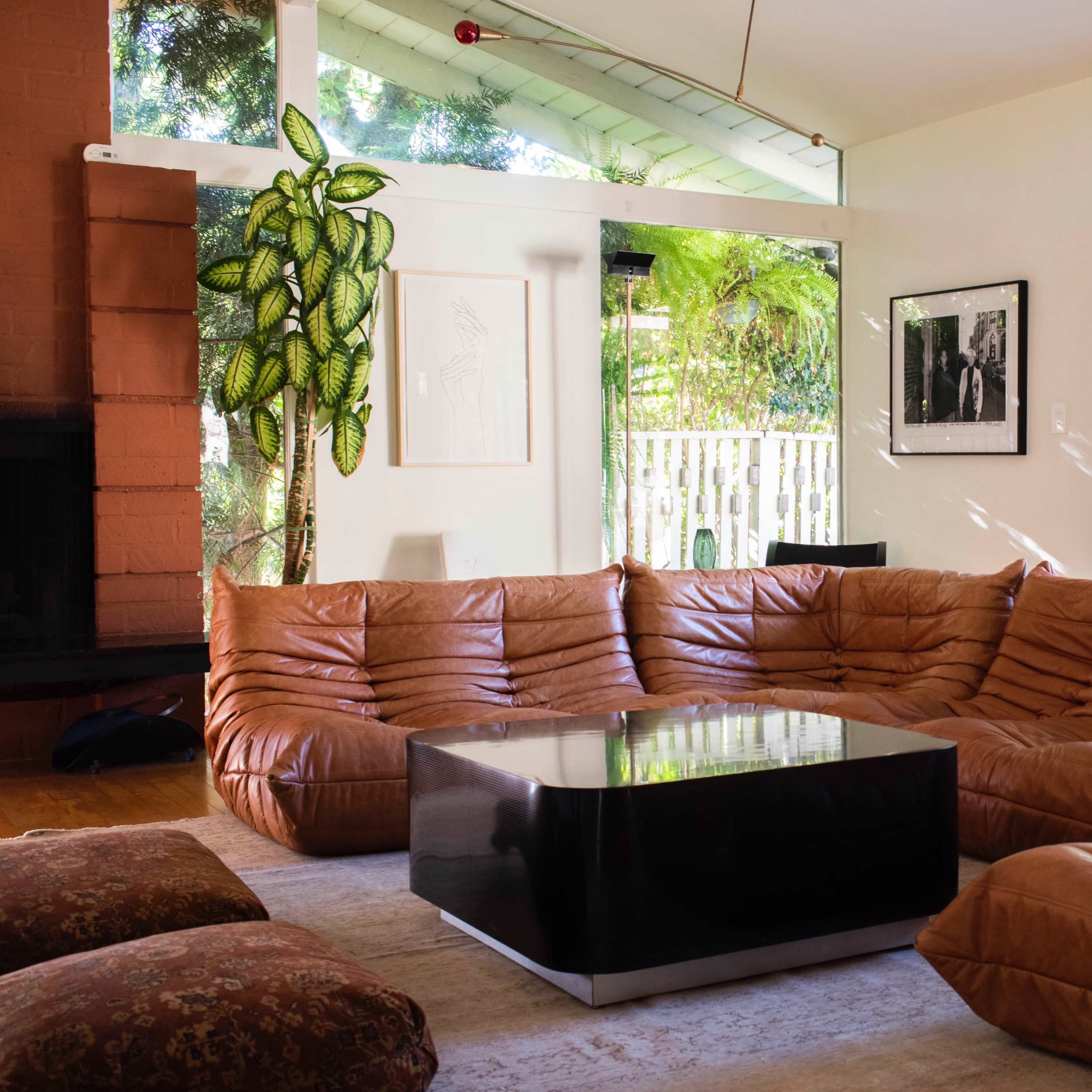 A spacious living room features a brown leather sectional sofa, a black coffee table, and large windows with views of greenery outside.