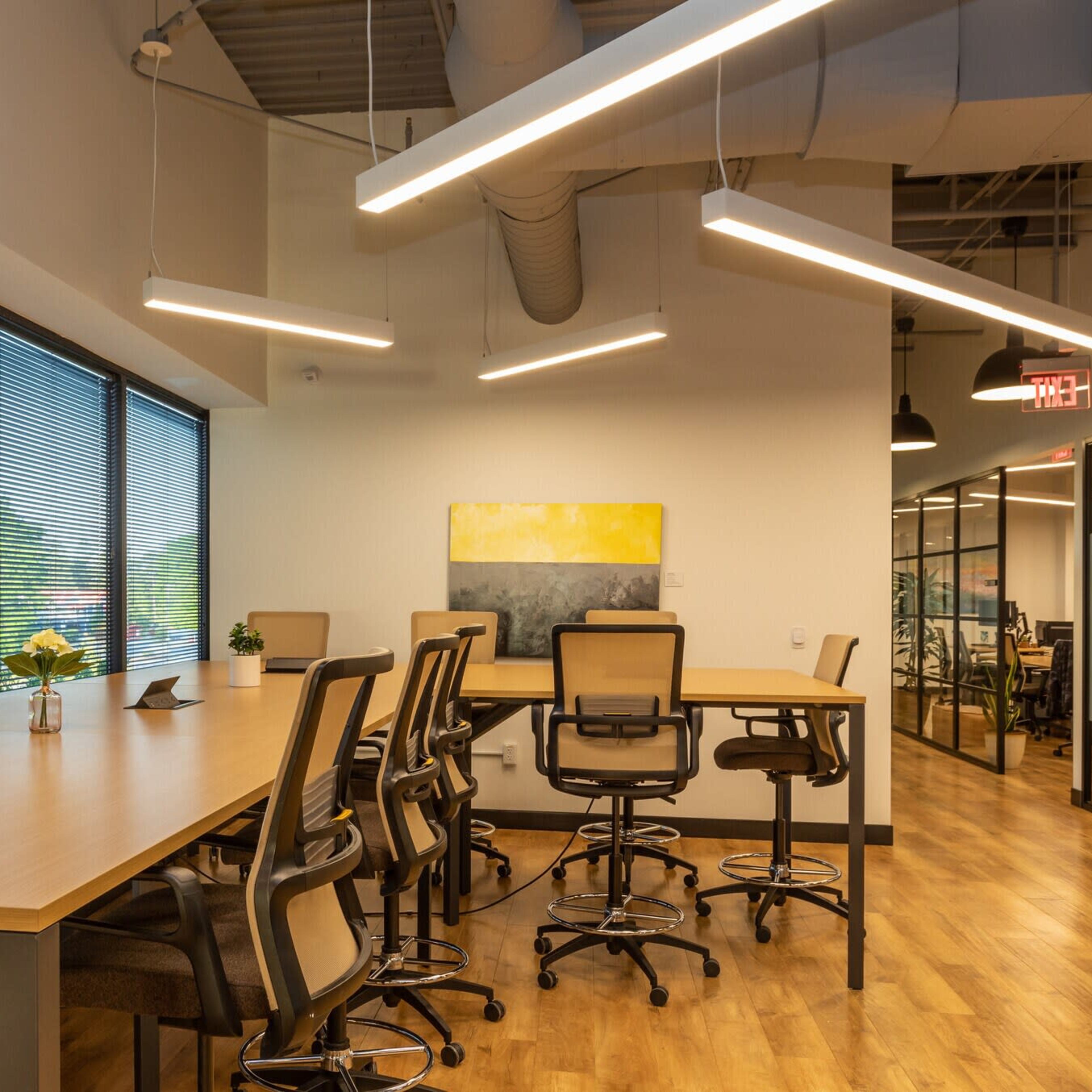 A modern office space with a long wooden table surrounded by high-backed chairs, large windows, and an adjacent glass-walled meeting area.