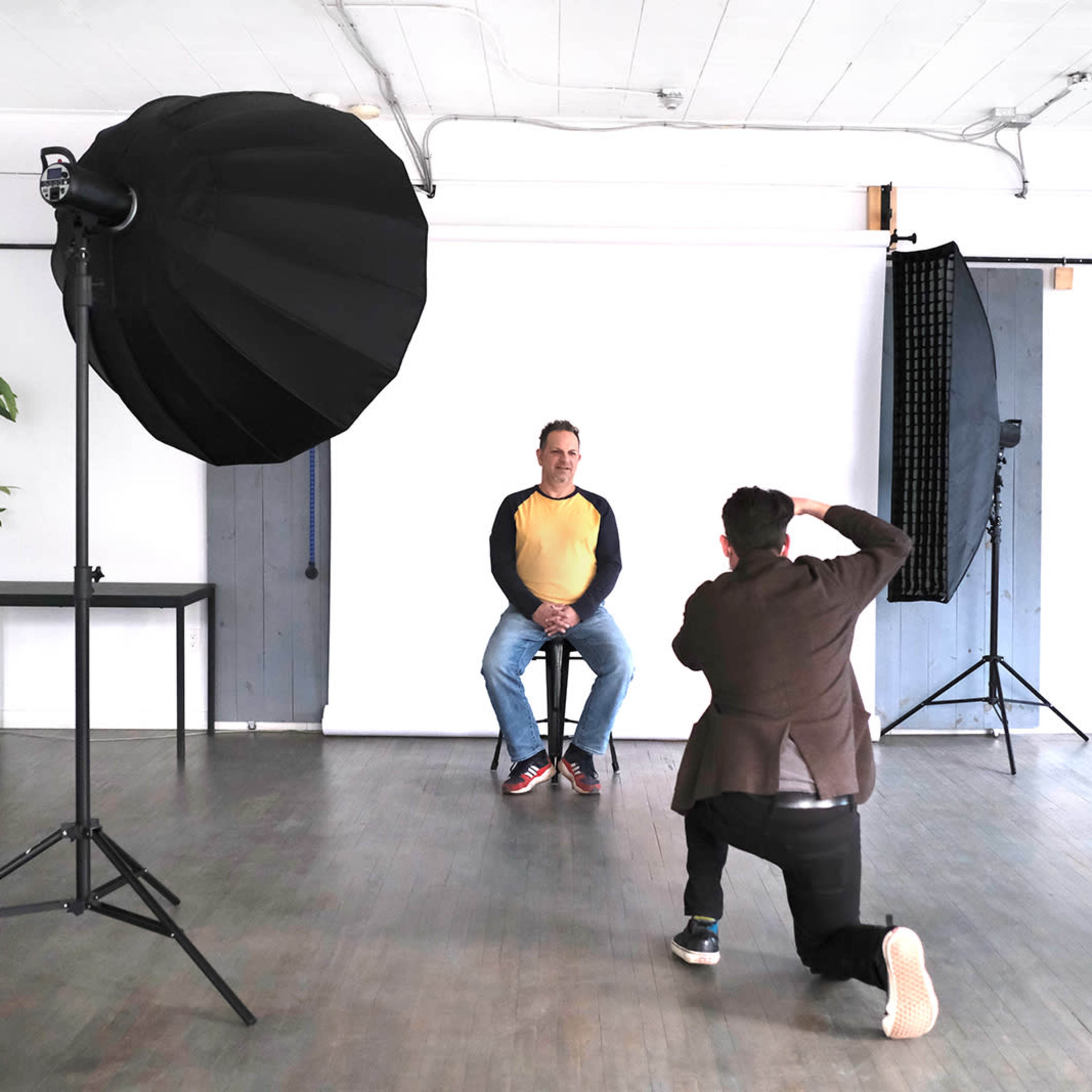 A photographer is capturing a portrait of a man sitting on a stool in a studio with a white backdrop and lighting equipment.