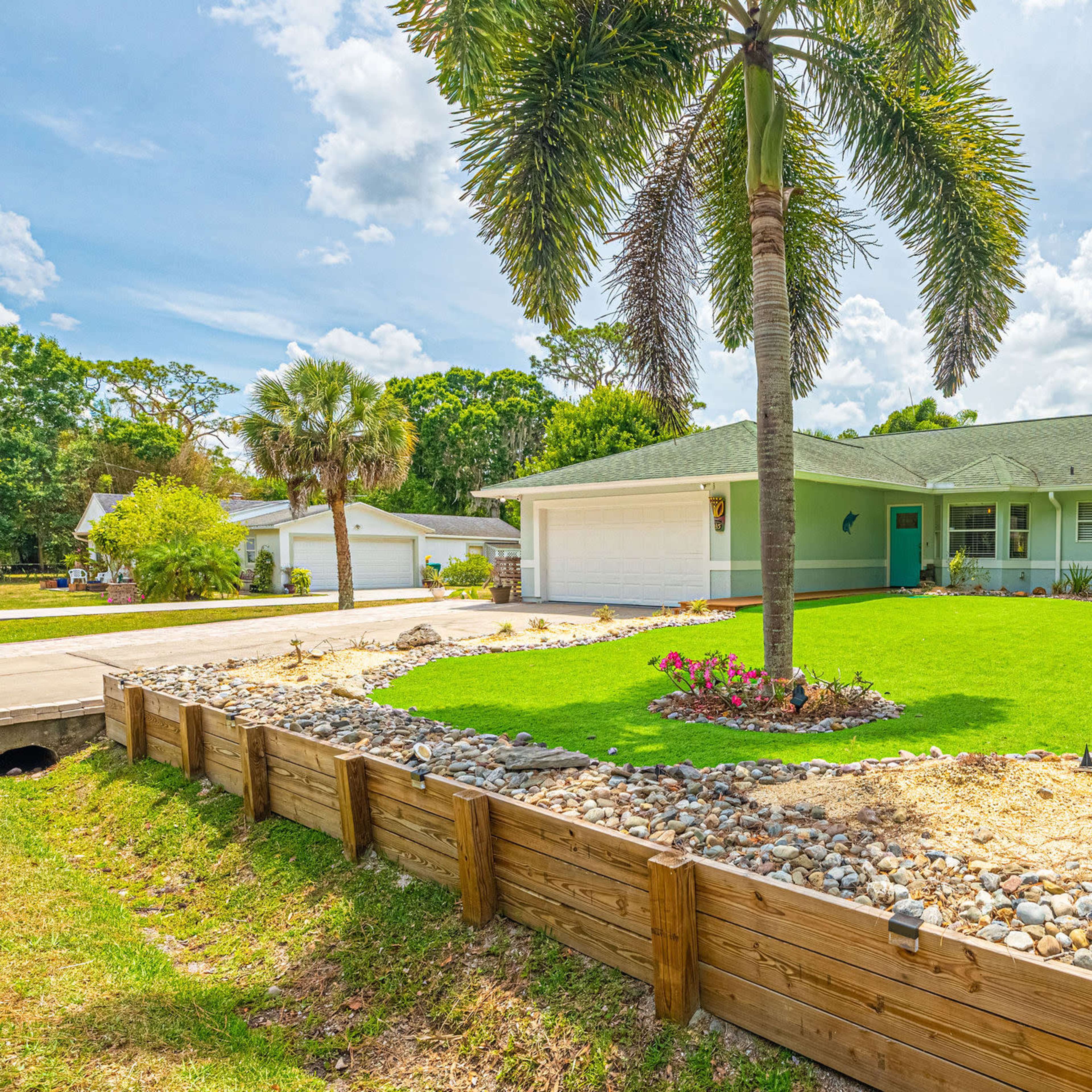 A residential property featuring a well-maintained lawn bordered by a wooden retaining wall and decorative stone landscaping, with a palm tree in the front yard and a two-car garage.
