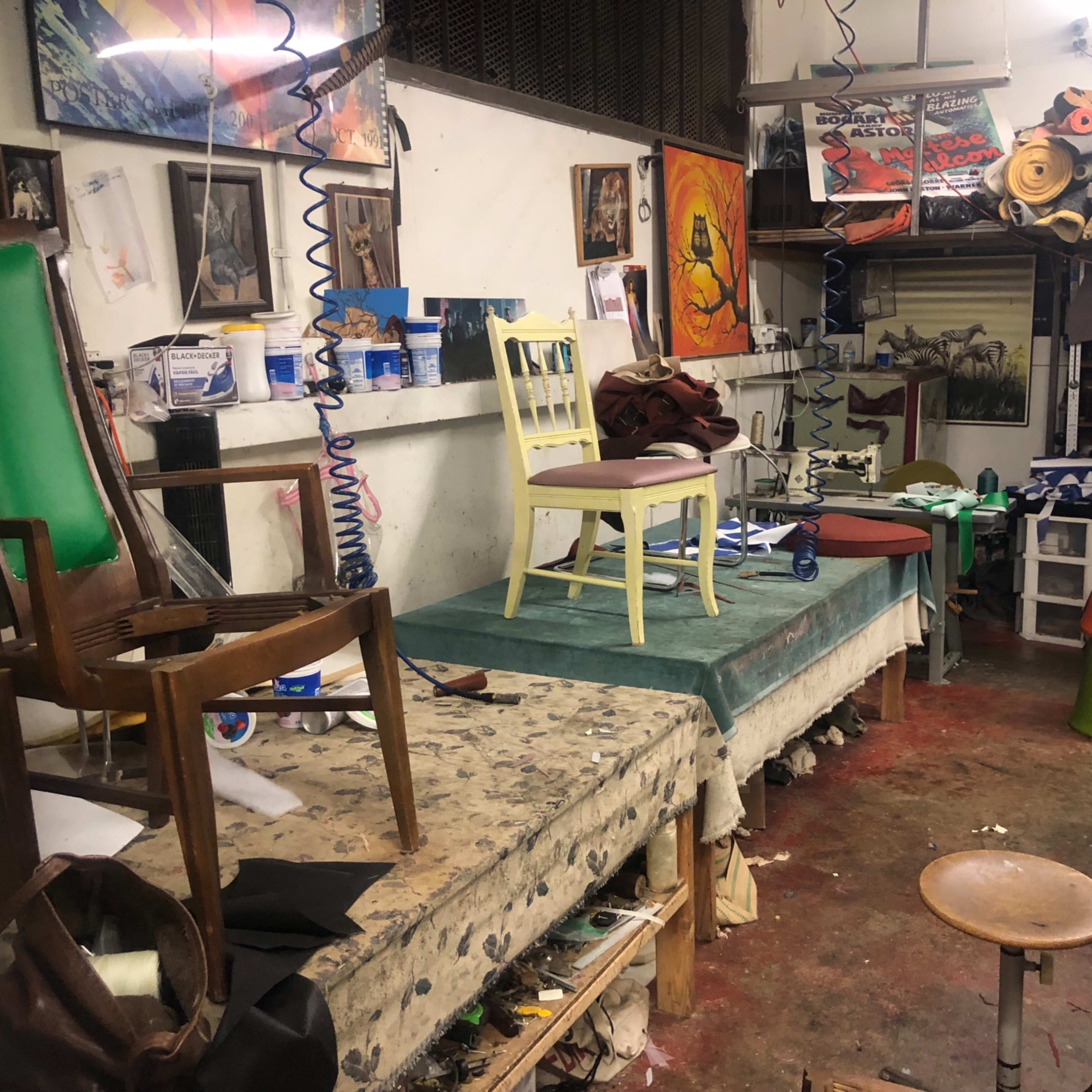 A cluttered workshop filled with various chairs, paint cans, and art supplies on tables, along with paintings on the walls.
