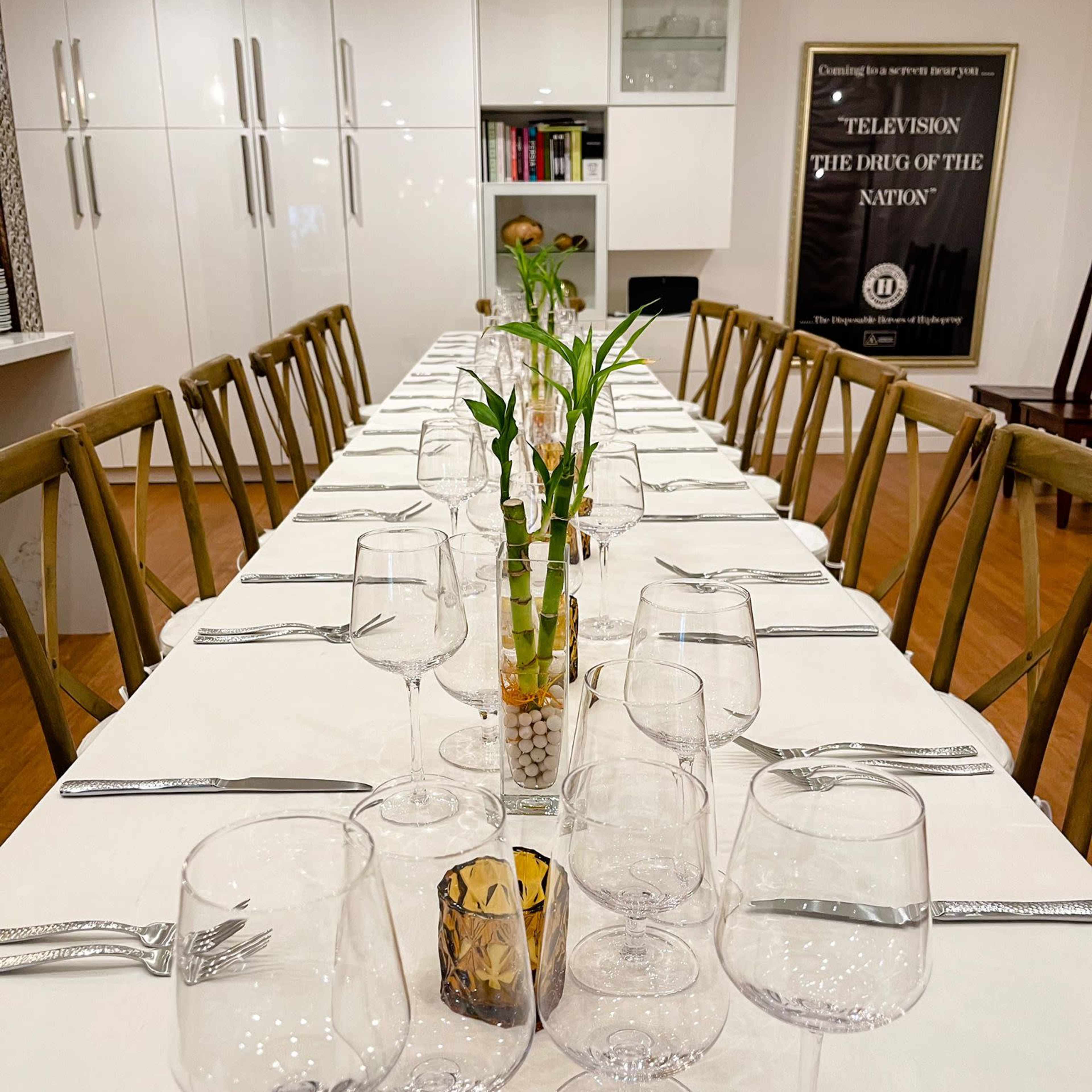 A long dining table is set with wine glasses, utensils, and a small decorative plant in the center, against a backdrop of a modern kitchen and a framed poster.