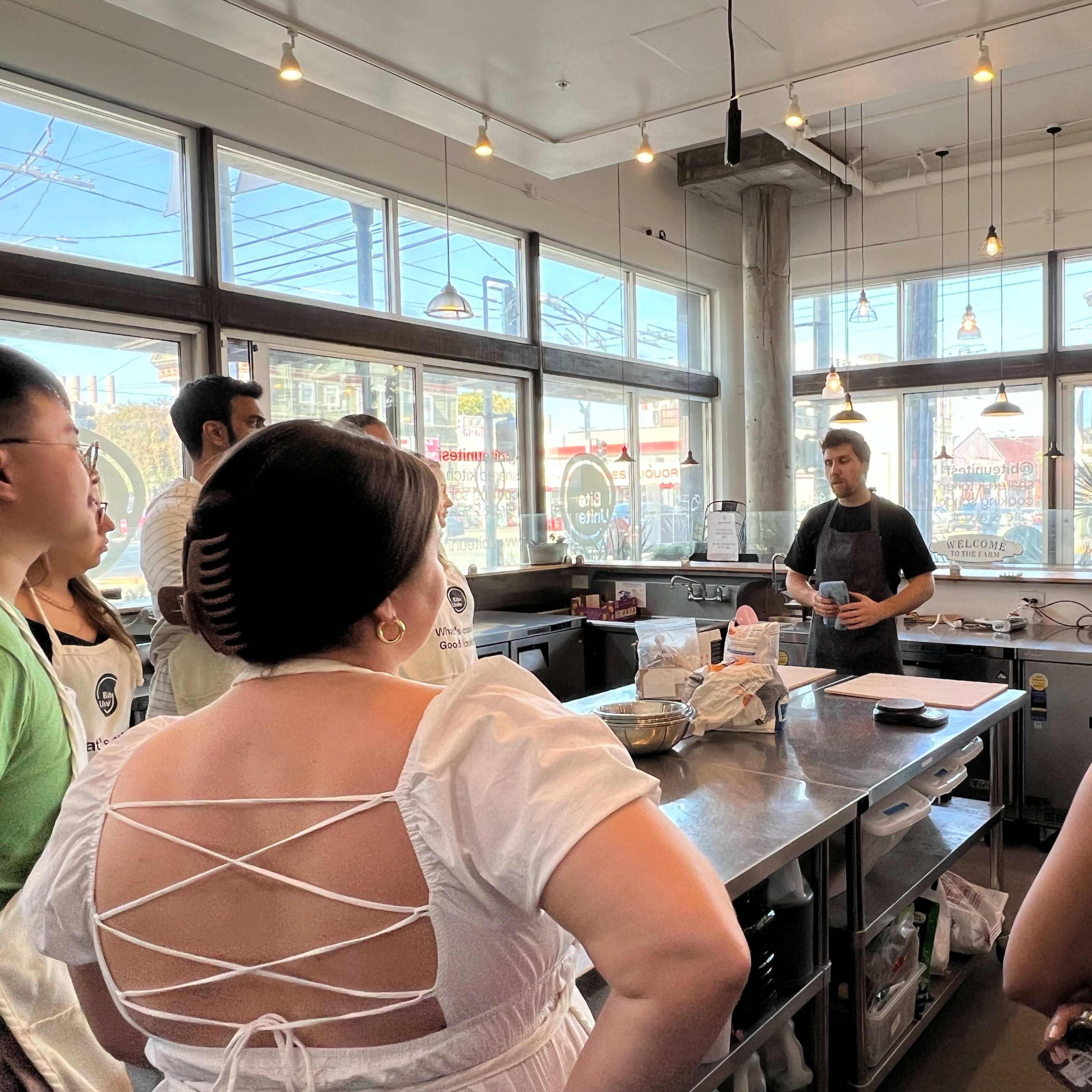 A group of people gathers around a chef instructing them in a brightly lit kitchen with large windows.