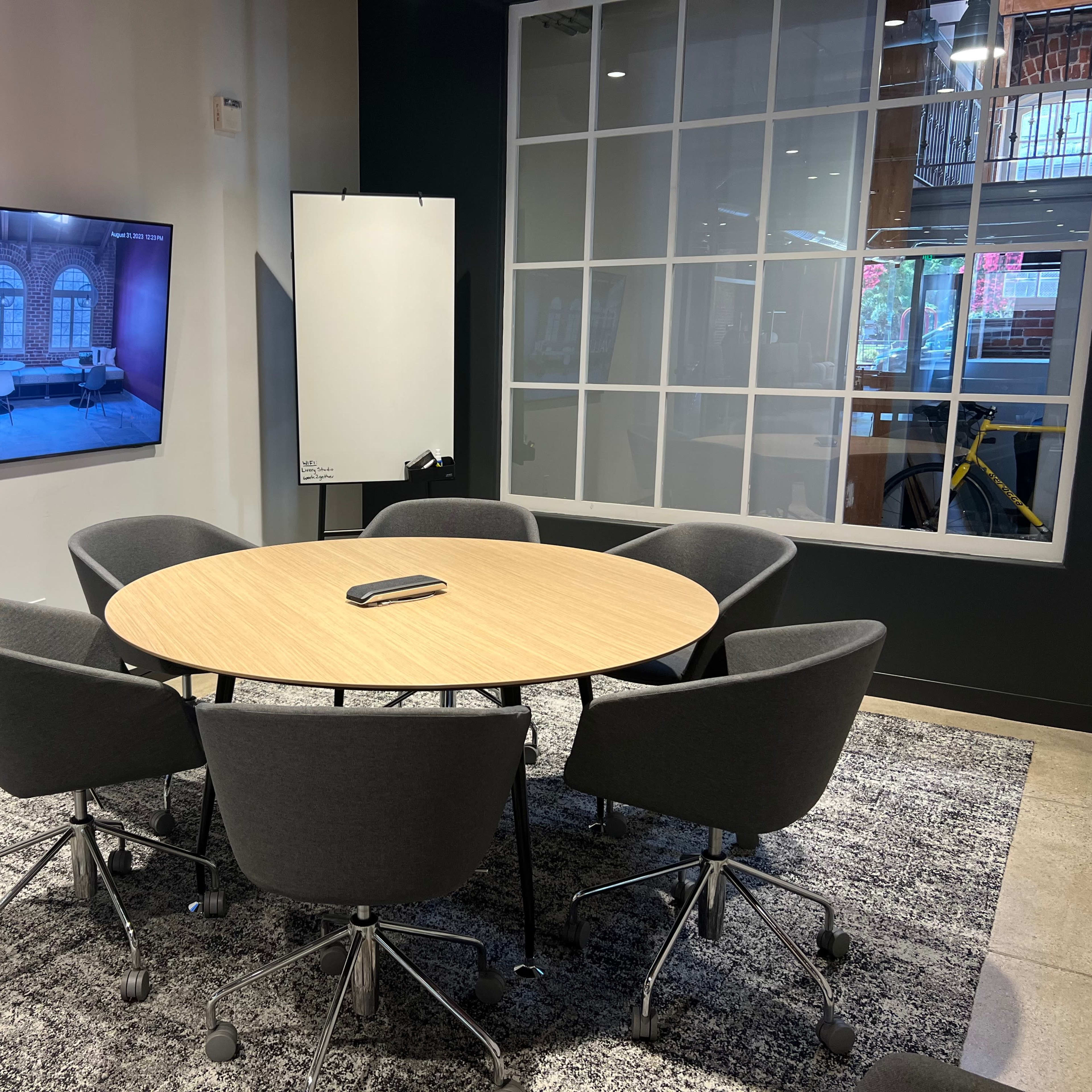 A modern conference room with a round table surrounded by six gray chairs, a large display screen on the wall, and a whiteboard.