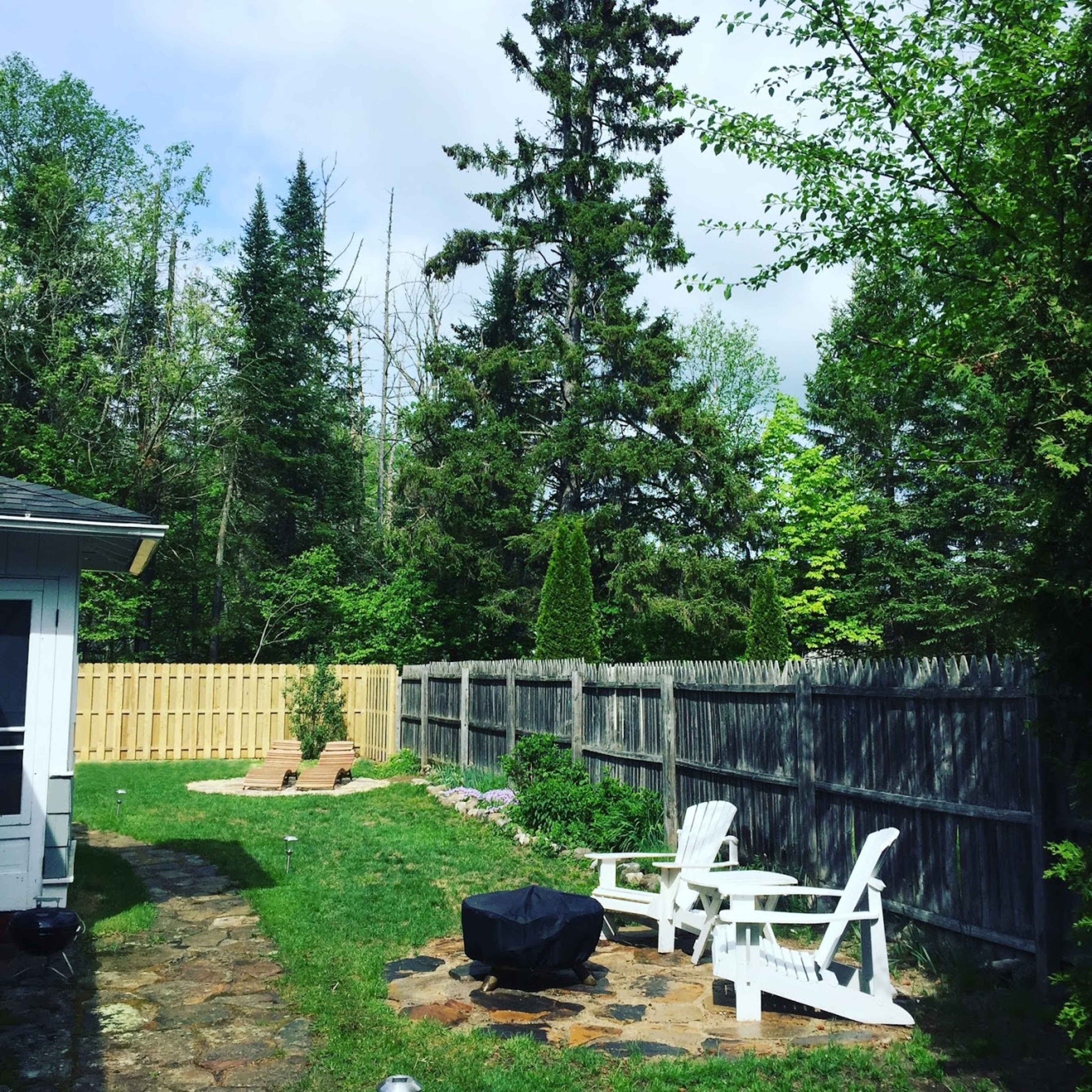 A backyard features a stone patio with two white chairs, a charcoal grill, and a wooden fence surrounded by trees and grass.