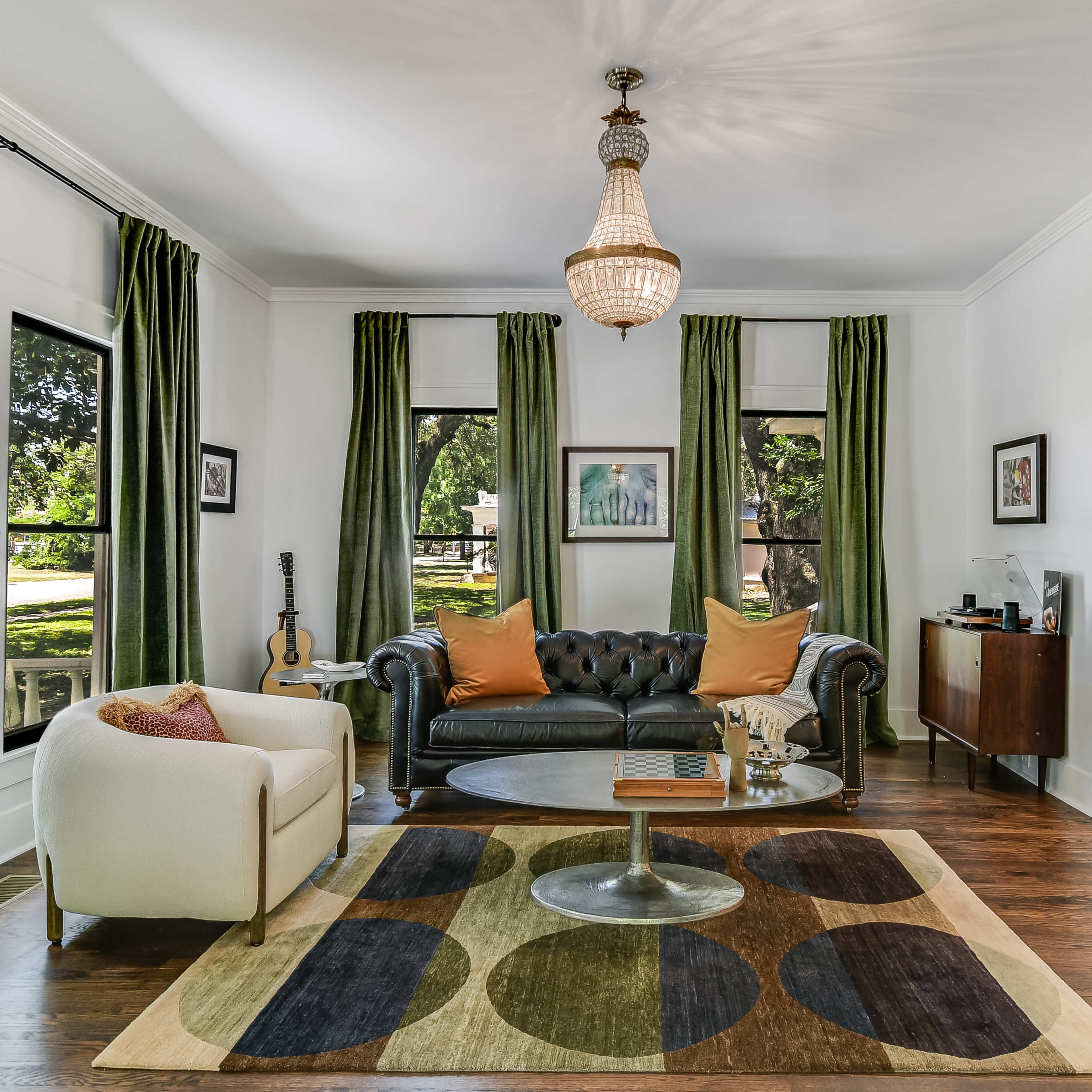 A living room features a black leather sofa, a white armchair, a circular coffee table, and large windows draped with green curtains.