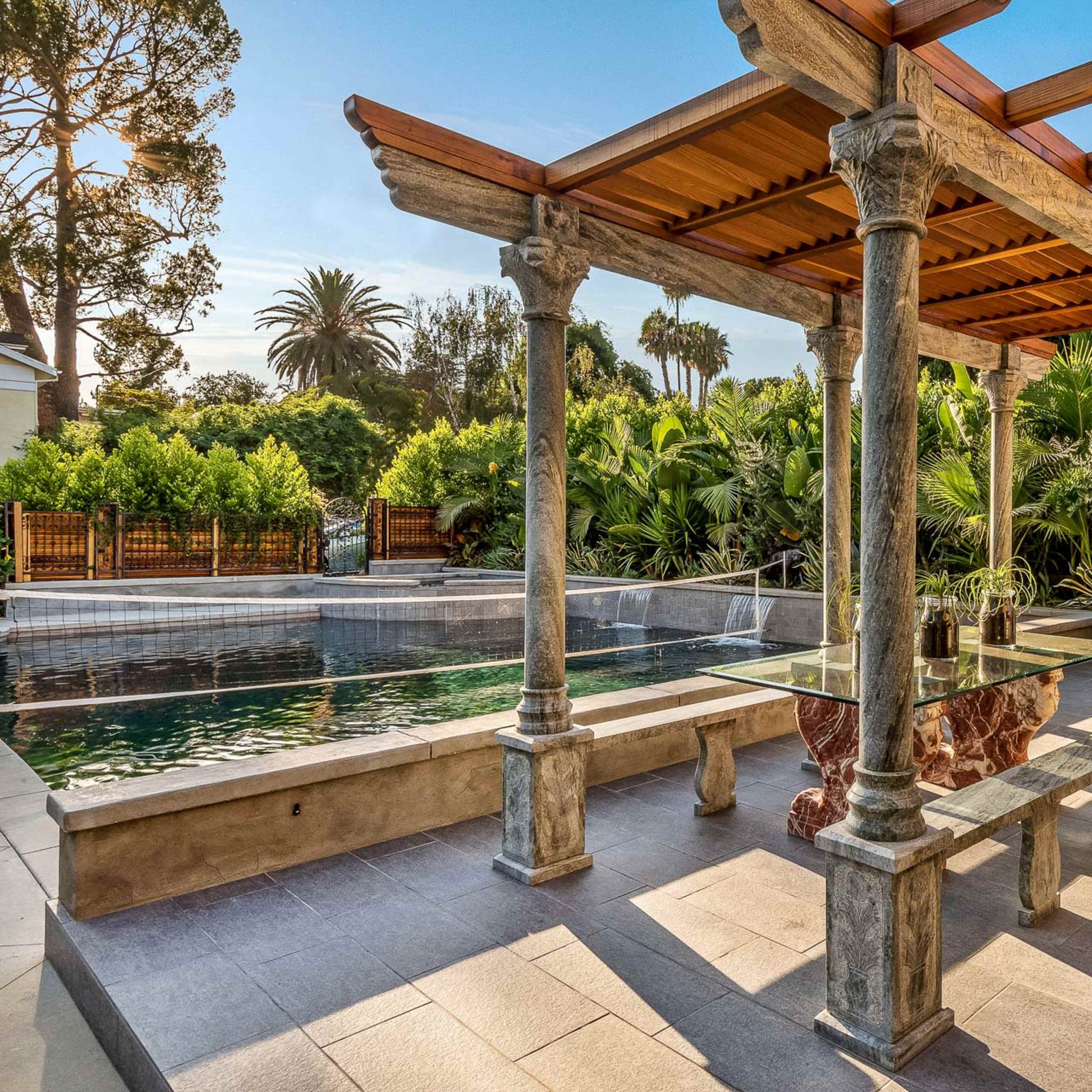 A patio area with a stone pergola, a swimming pool, and lush greenery surrounding the space.