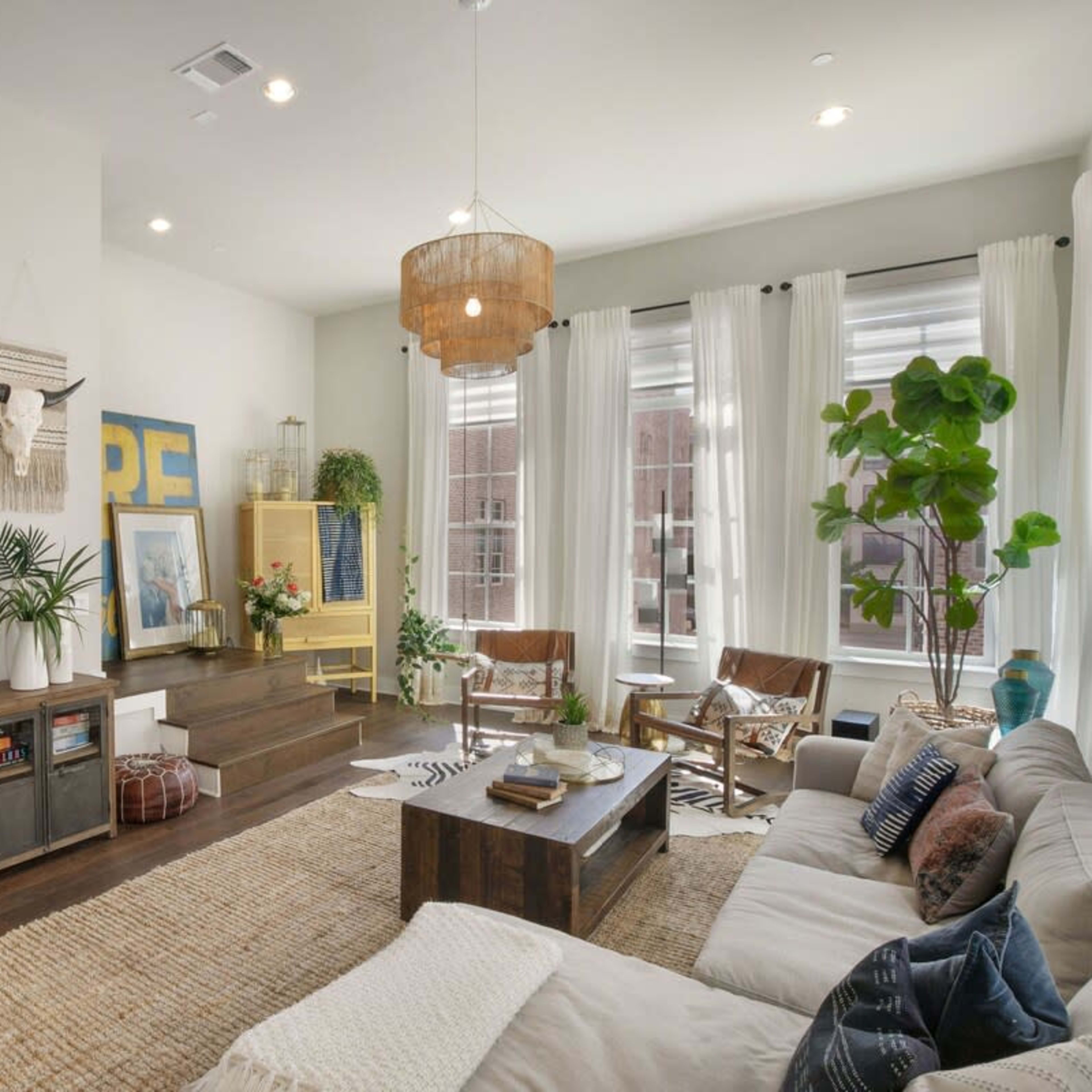 The living room features a flat-screen TV on the wall, a large coffee table, and a mix of seating including a sectional sofa and wooden chairs, all arranged around a neutral-colored area rug.