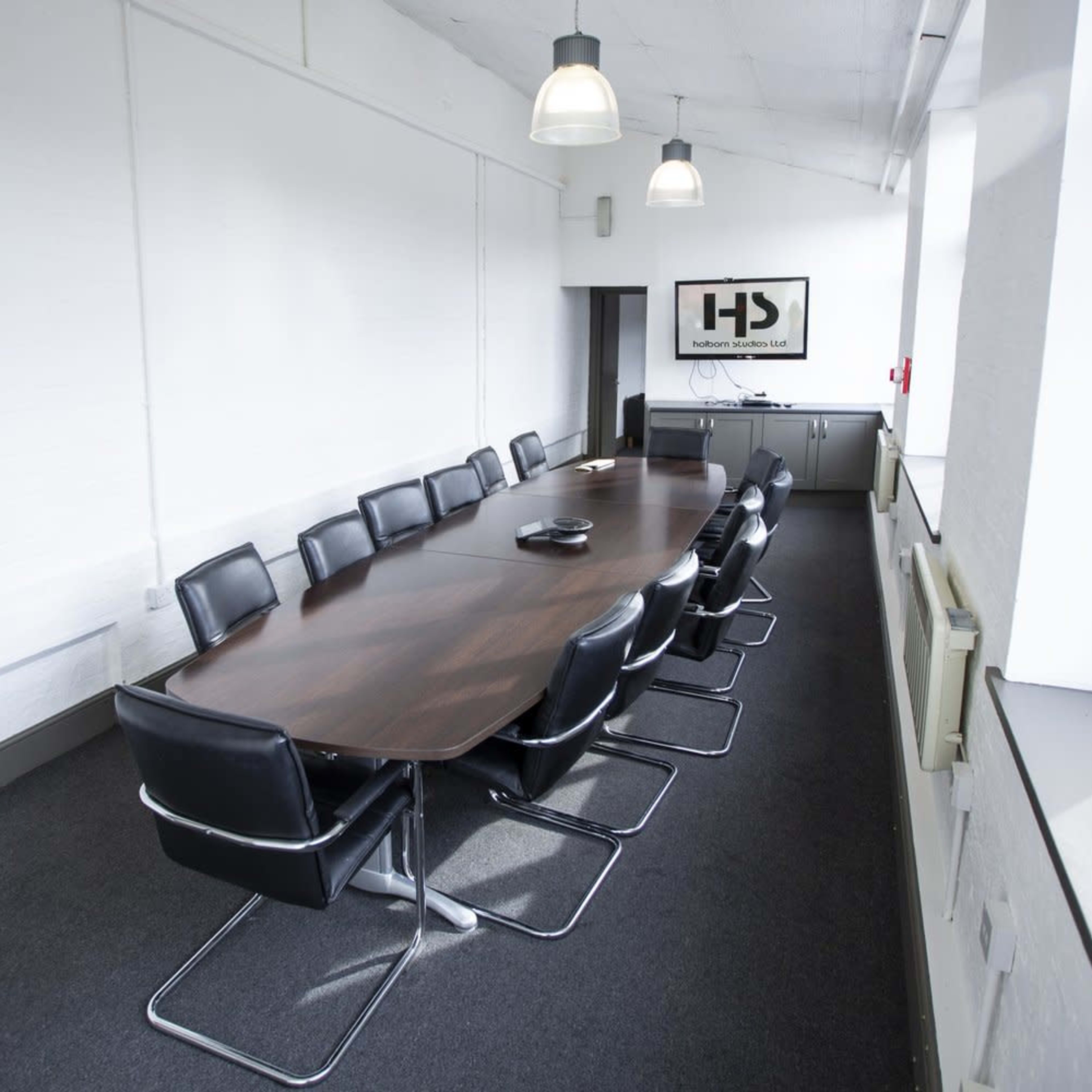 A long conference table with black chairs sits in a bright, modern meeting room featuring large windows and a wall-mounted screen.