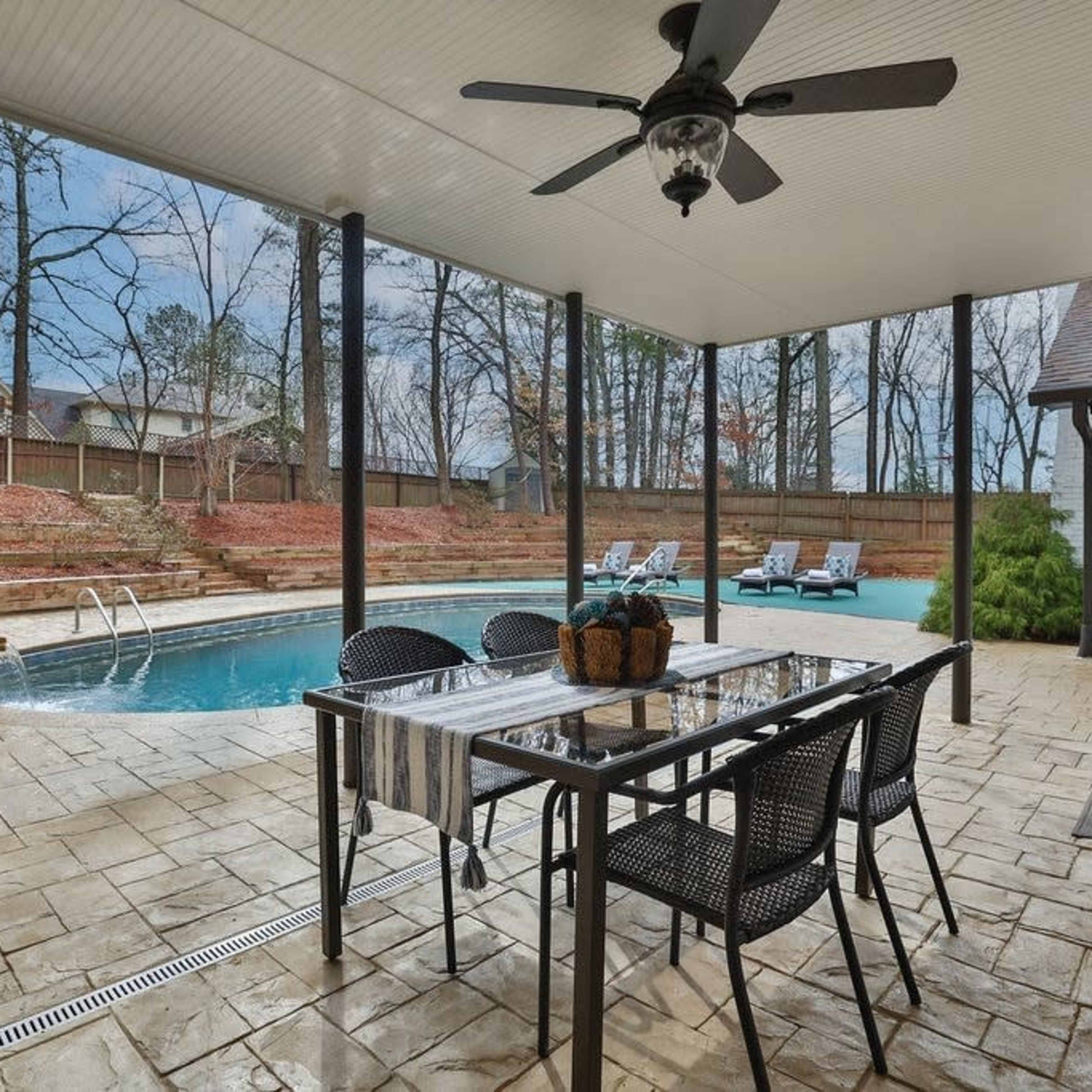 A covered outdoor patio with a dining table overlooks a swimming pool surrounded by stone, set in a wooded area.