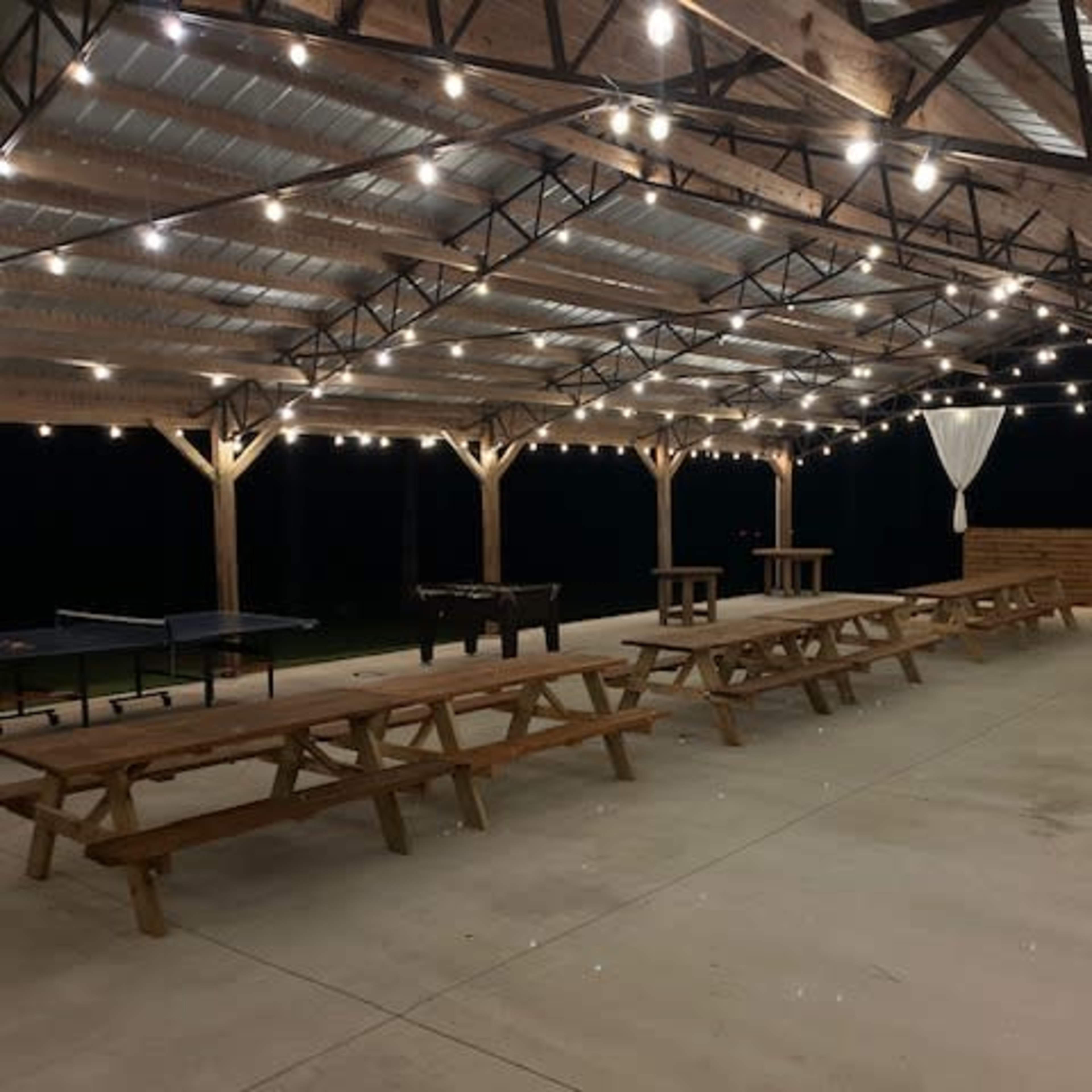 An outdoor pavilion with string lights, wooden picnic tables, and a bar area, set against a dark background.