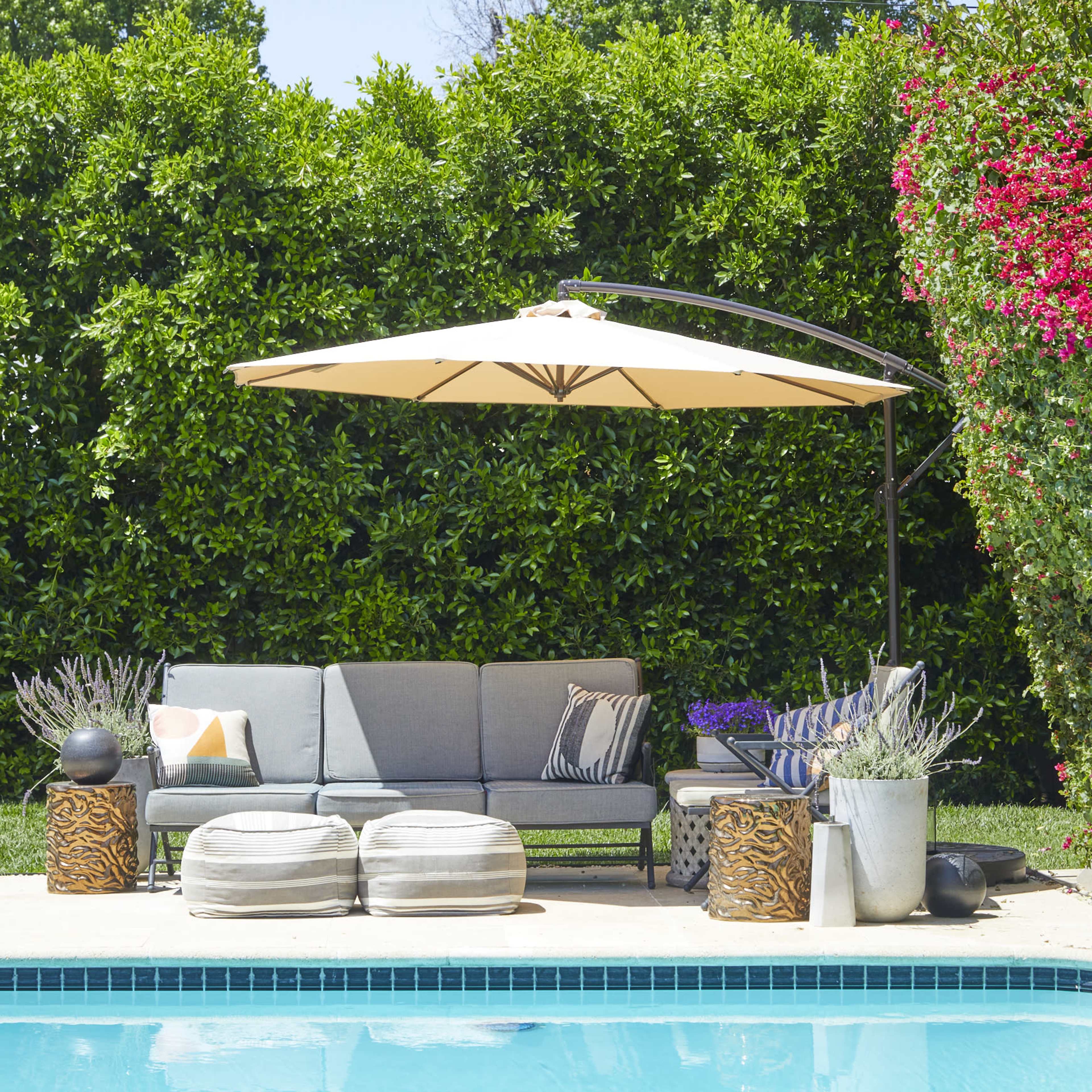 A modern outdoor seating area with a gray couch, large umbrella, decorative plants, and a pool in the foreground.