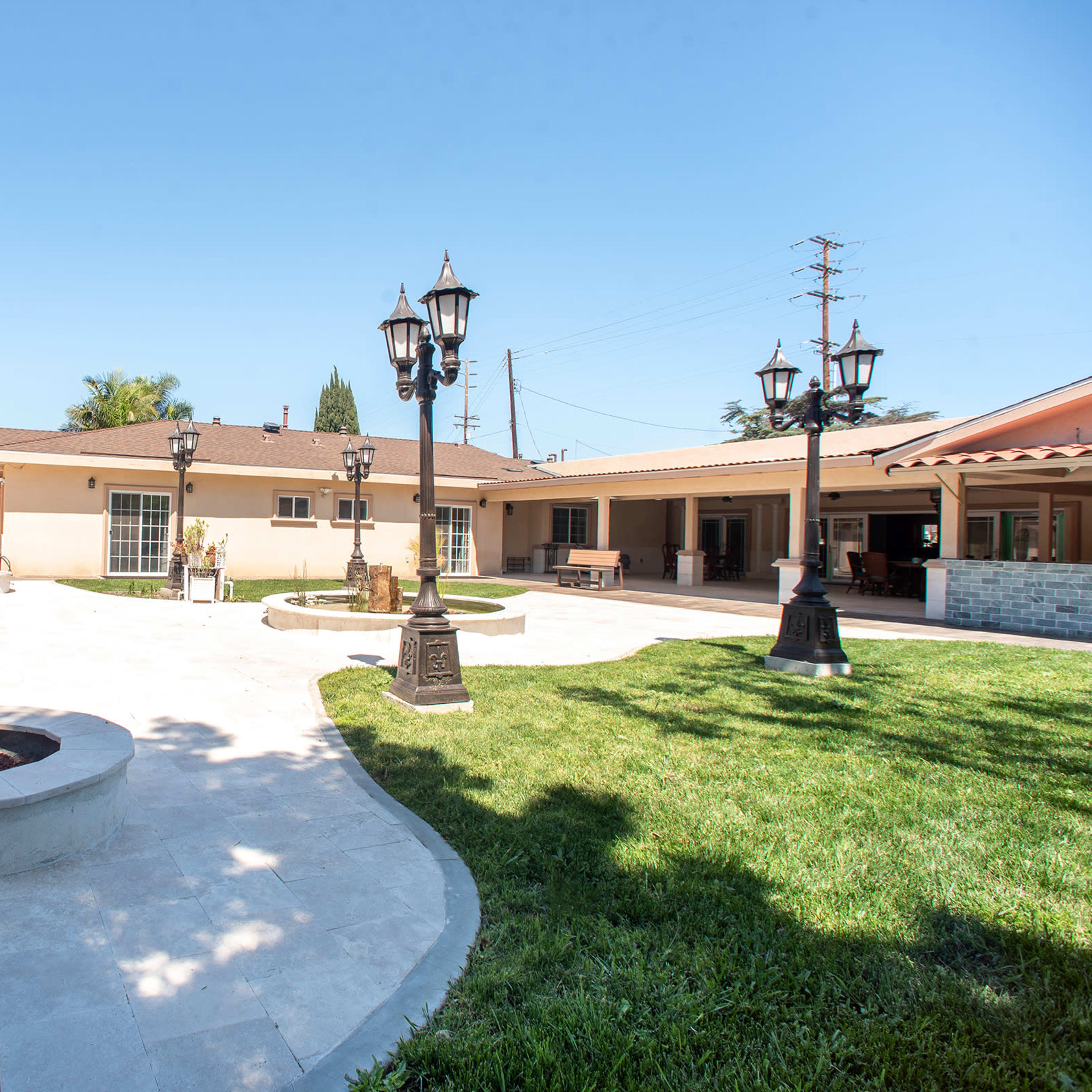 A spacious backyard with a paved area, circular fire pit, and multiple lantern-style lamps surrounding a lawn and patio area.
