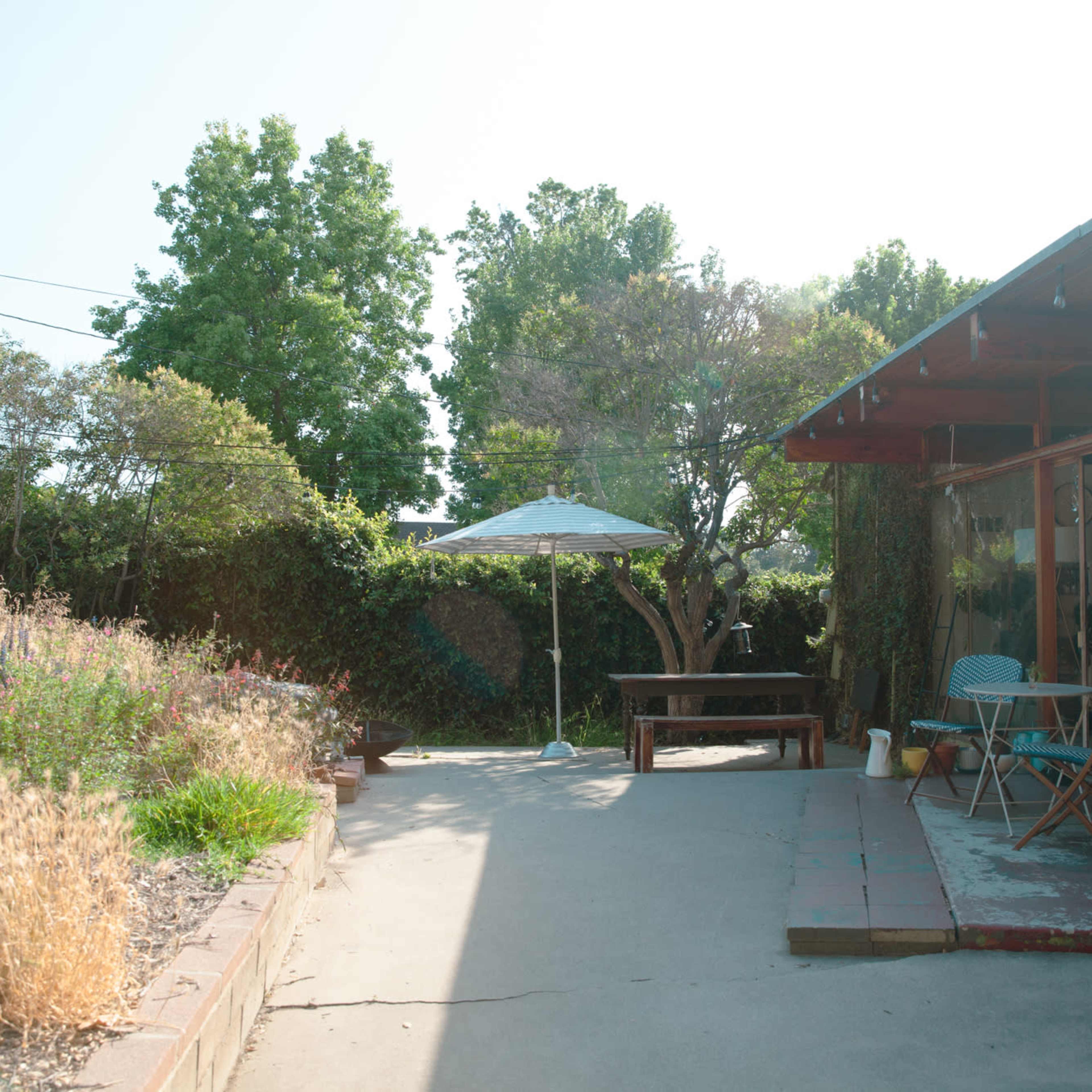 A shaded patio area with a table and chairs is surrounded by a garden of tall grass and trees.