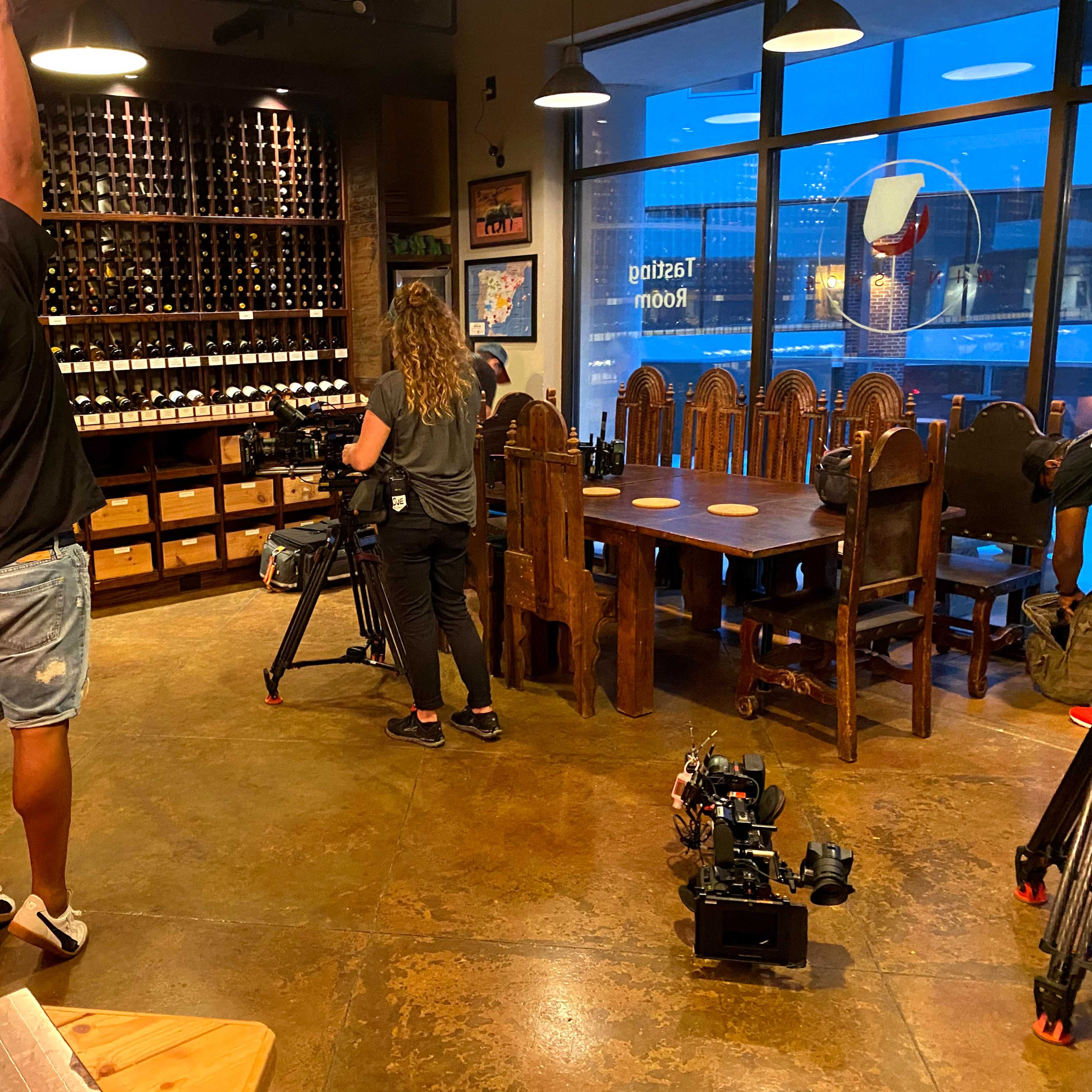A man adjusts a light fixture while a woman sets up a camera in a restaurant with wooden furniture and a wall of wine bottles.