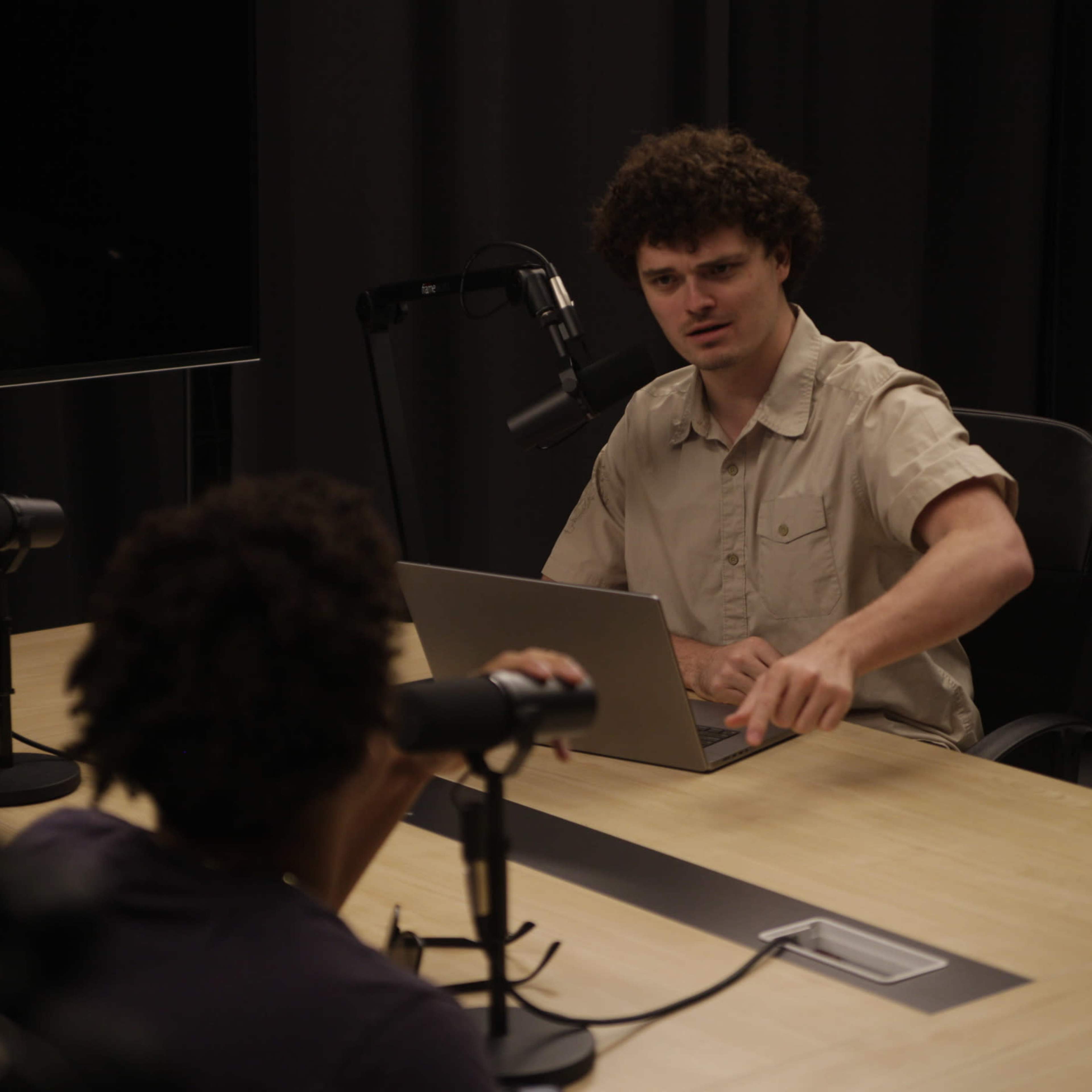 Two individuals are seated at a table with a laptop and microphones, engaged in a discussion in a dimly lit studio.