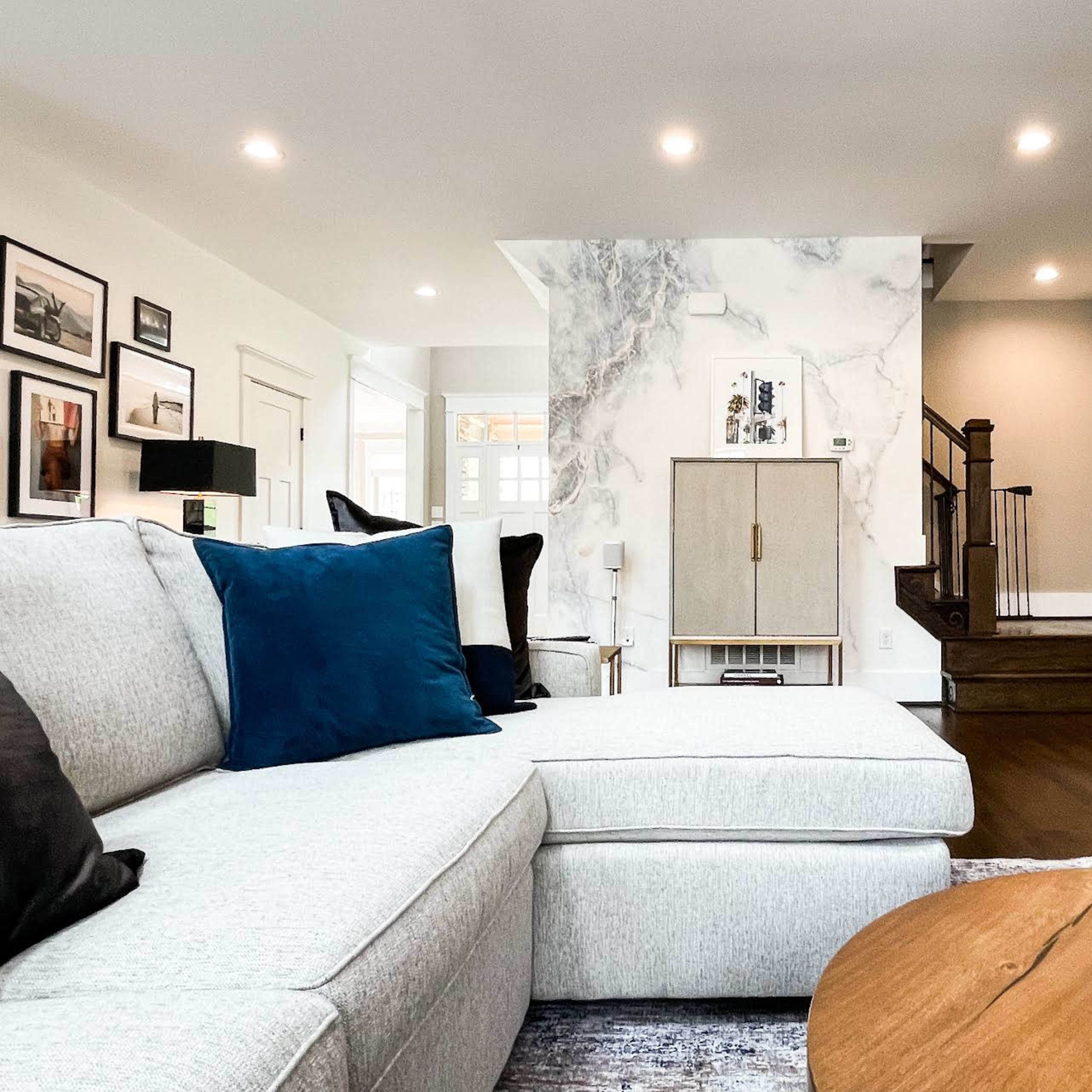 A modern living room with a light-colored sectional sofa, a wooden coffee table, and a staircase leading to another level, accented by a marble wall.