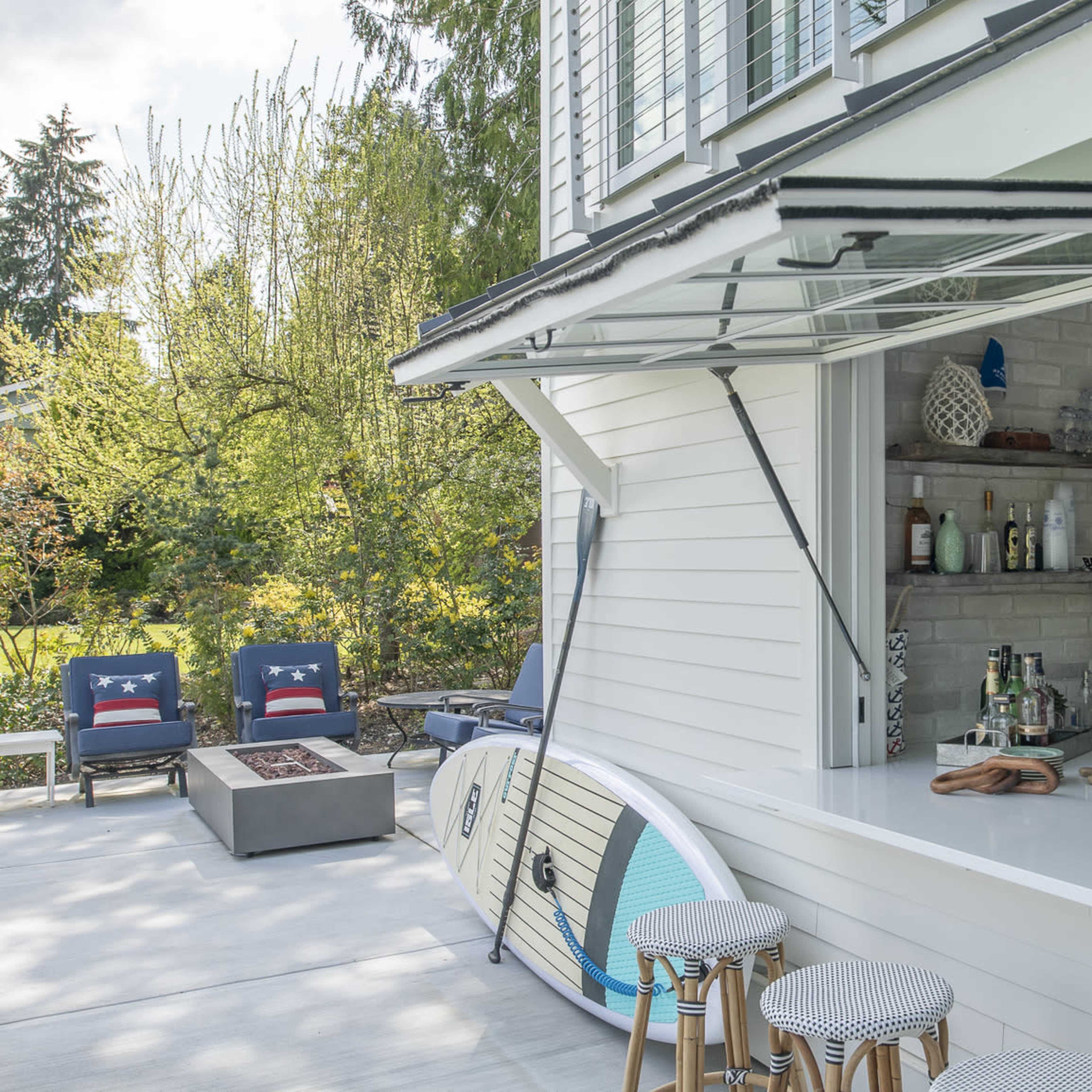 An outdoor bar area with a countertop, stools, and a shelf displaying various beverages, beside a paddleboard resting against the wall of a house.