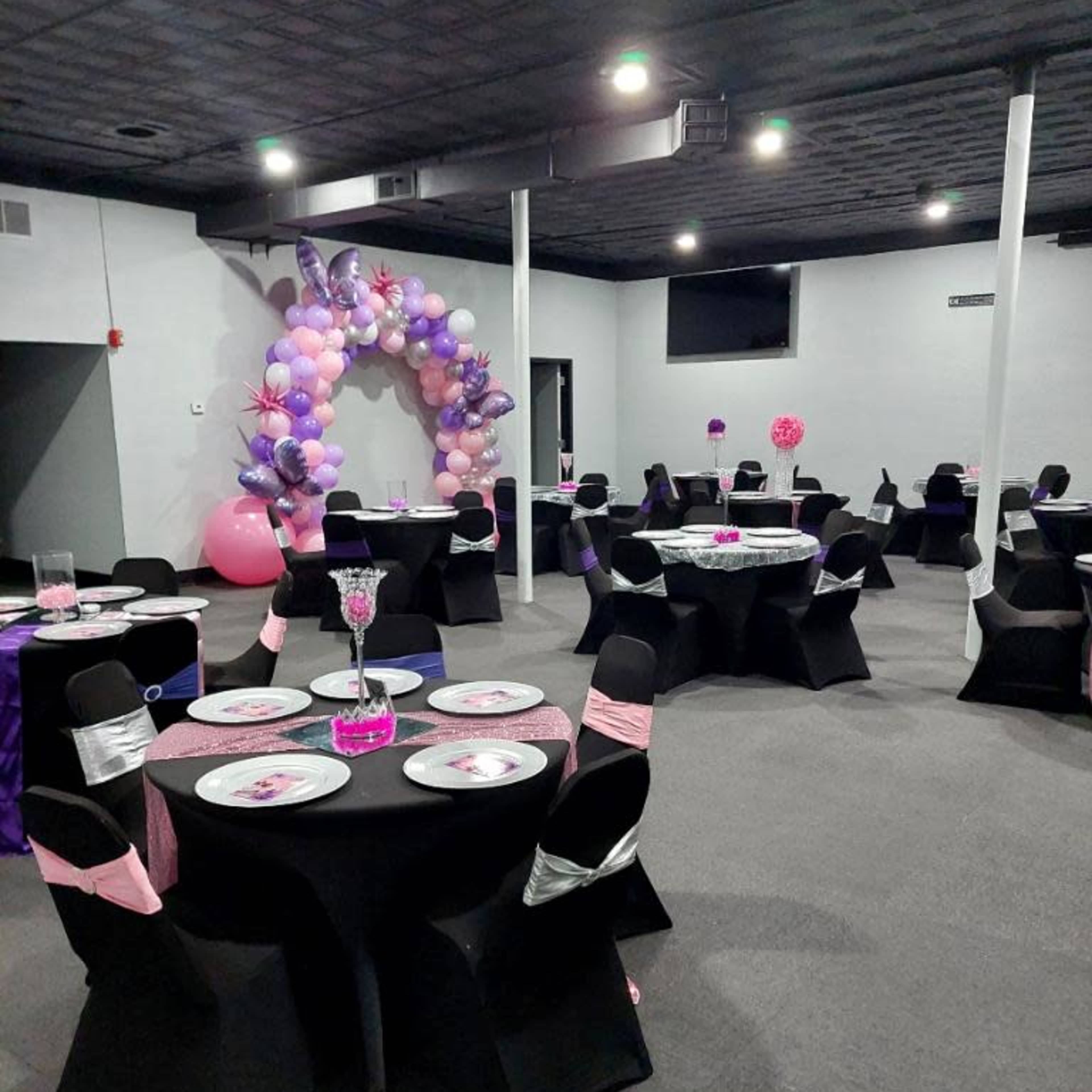 A party venue with black tables adorned with pink and purple decorations, surrounded by balloon arches and centerpieces.