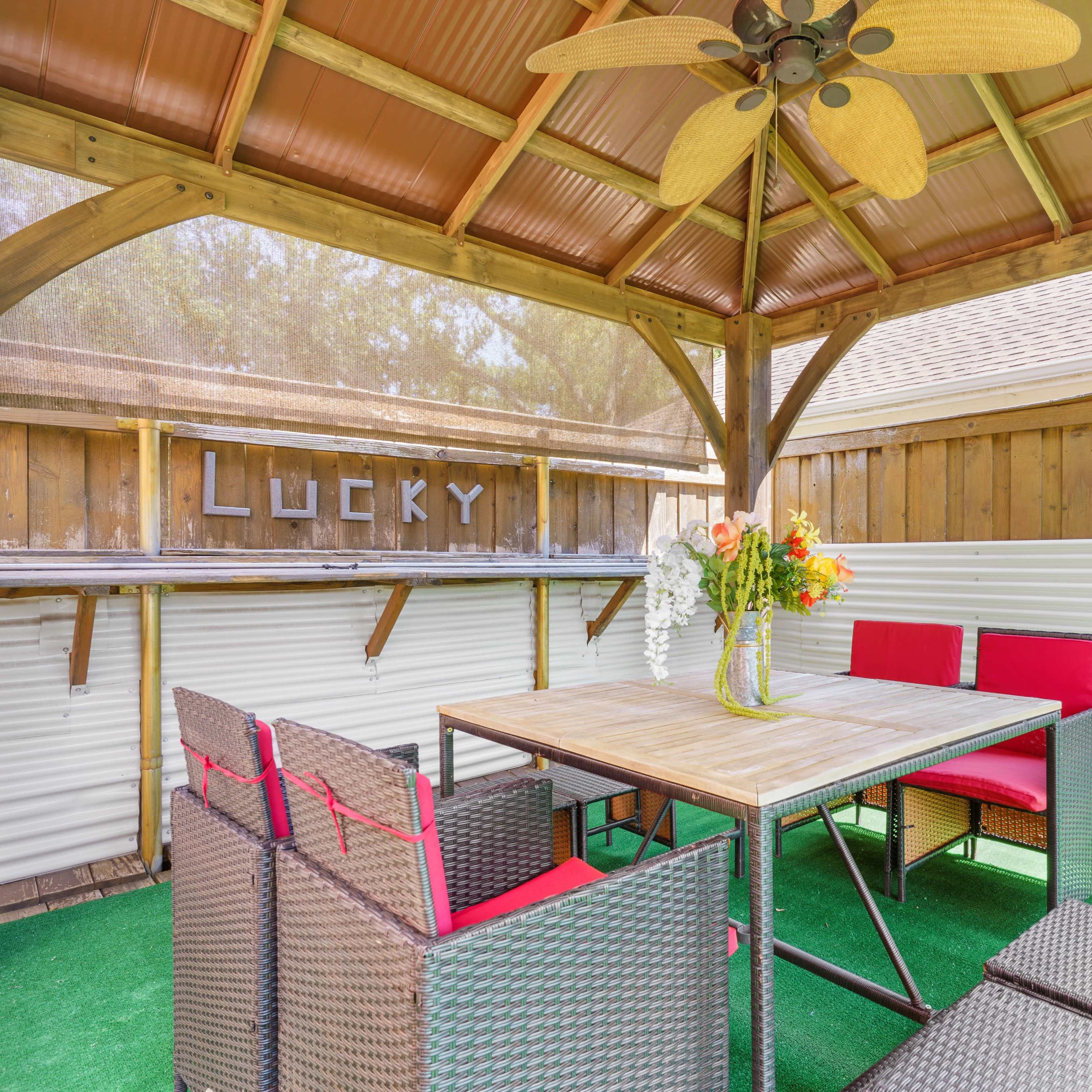 A shaded outdoor sitting area with a table, four red chairs, and a floral centerpiece under a wooden gazebo.