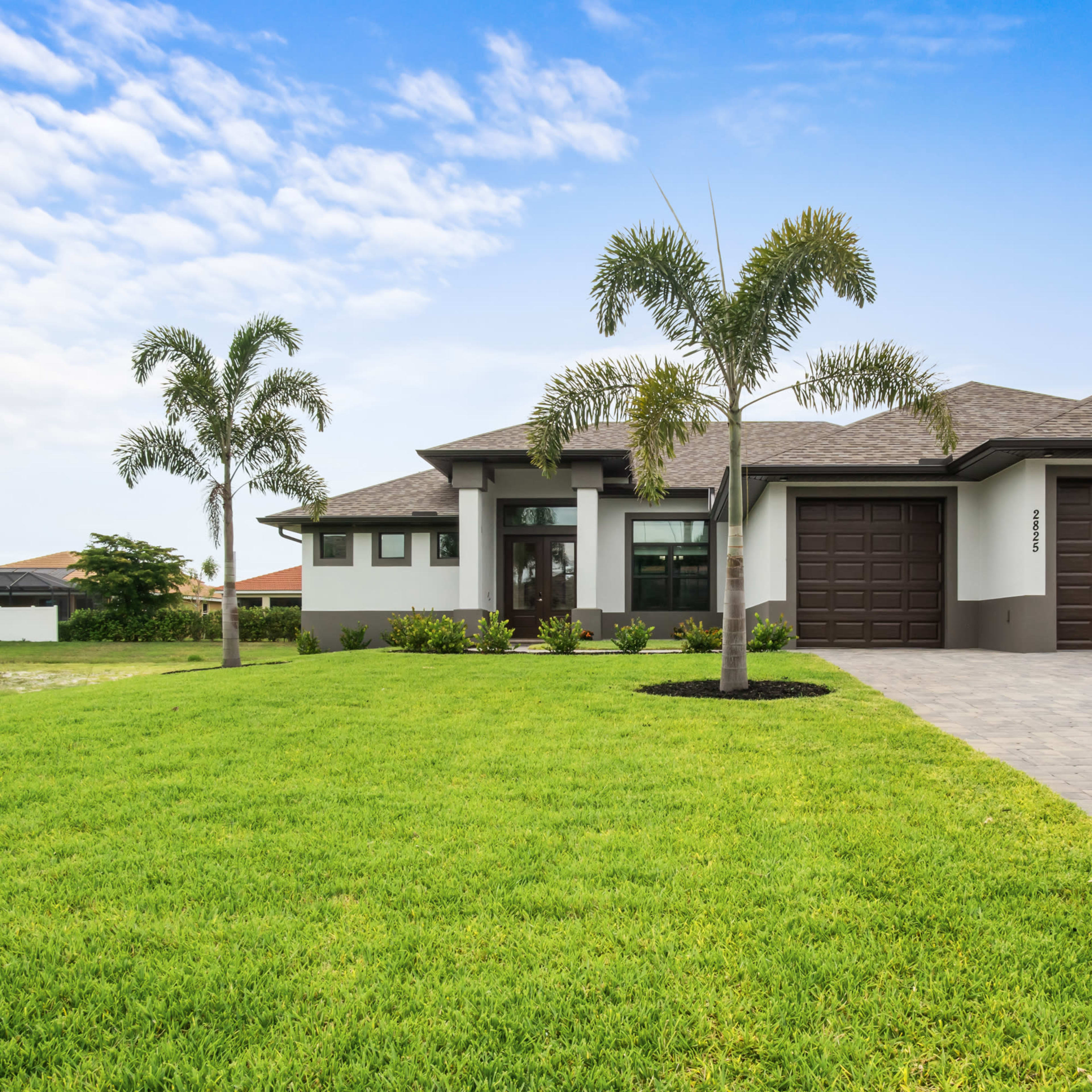 A modern single-story house with a two-car garage, surrounded by well-maintained grass and palm trees.