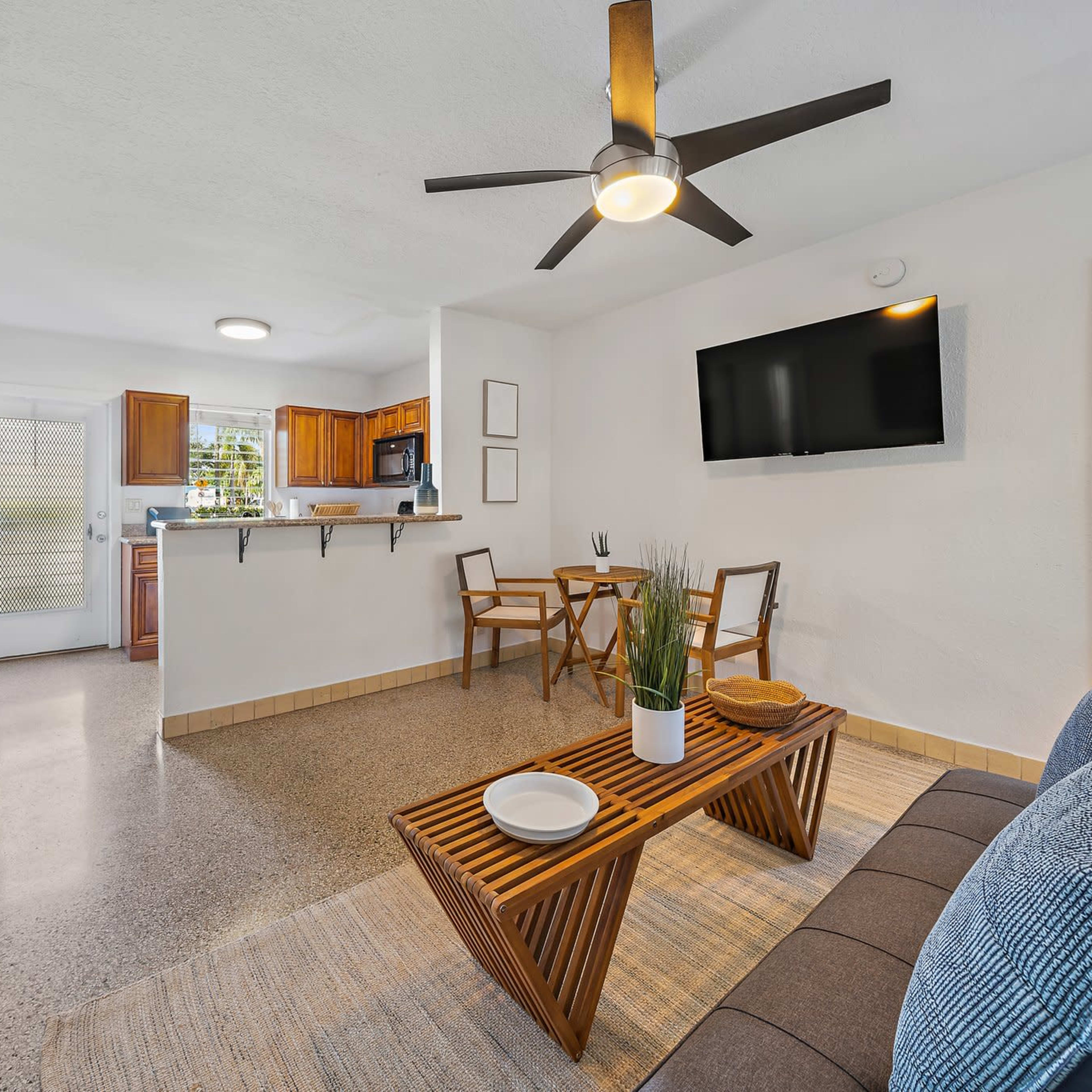 A modern open-concept living area featuring a seating area with a brown sofa, a wooden coffee table, a dining space with two chairs, and a kitchen in the background.