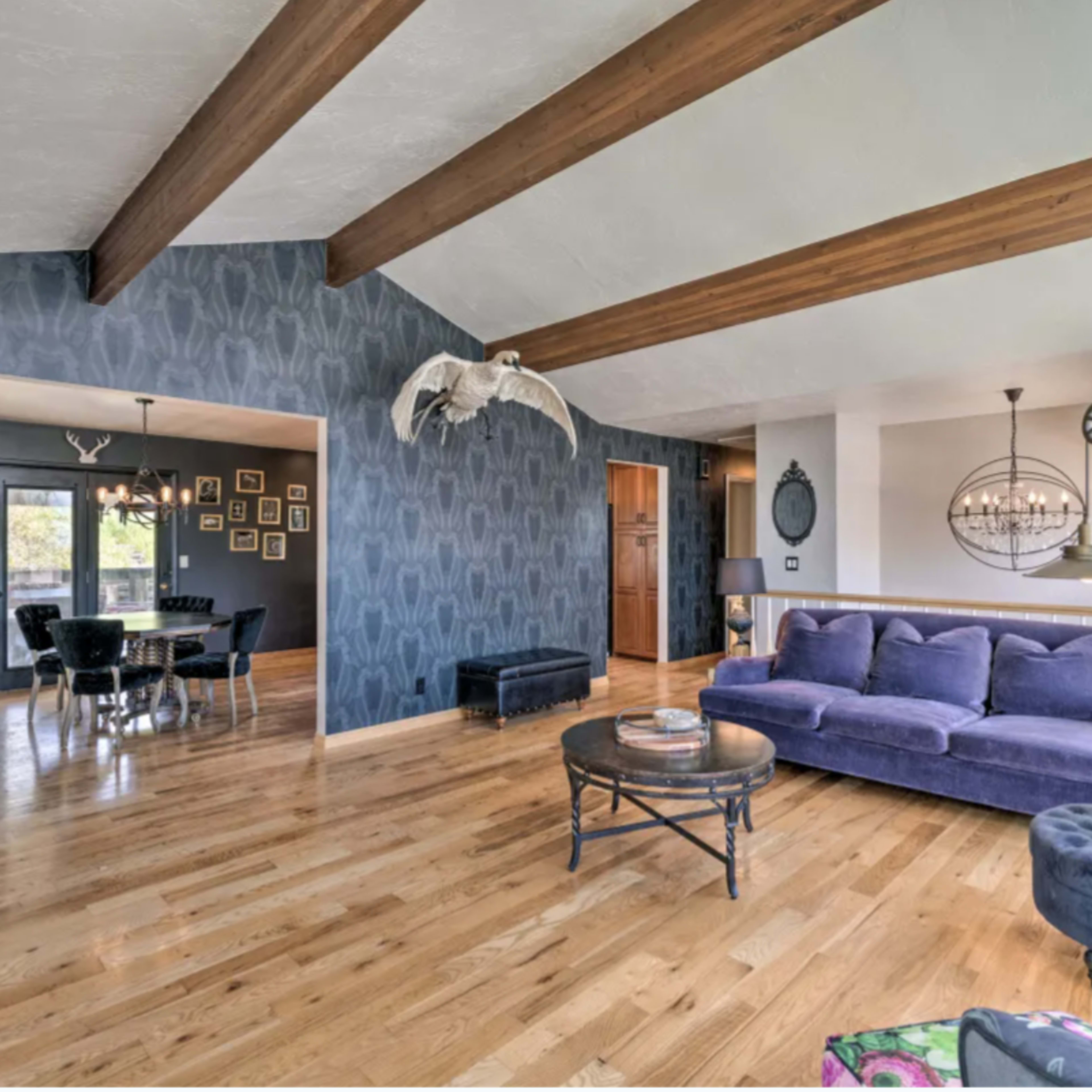 The spacious living room features hardwood floors, vaulted ceilings with exposed wooden beams, a purple sofa, and large windows that offer natural light.
