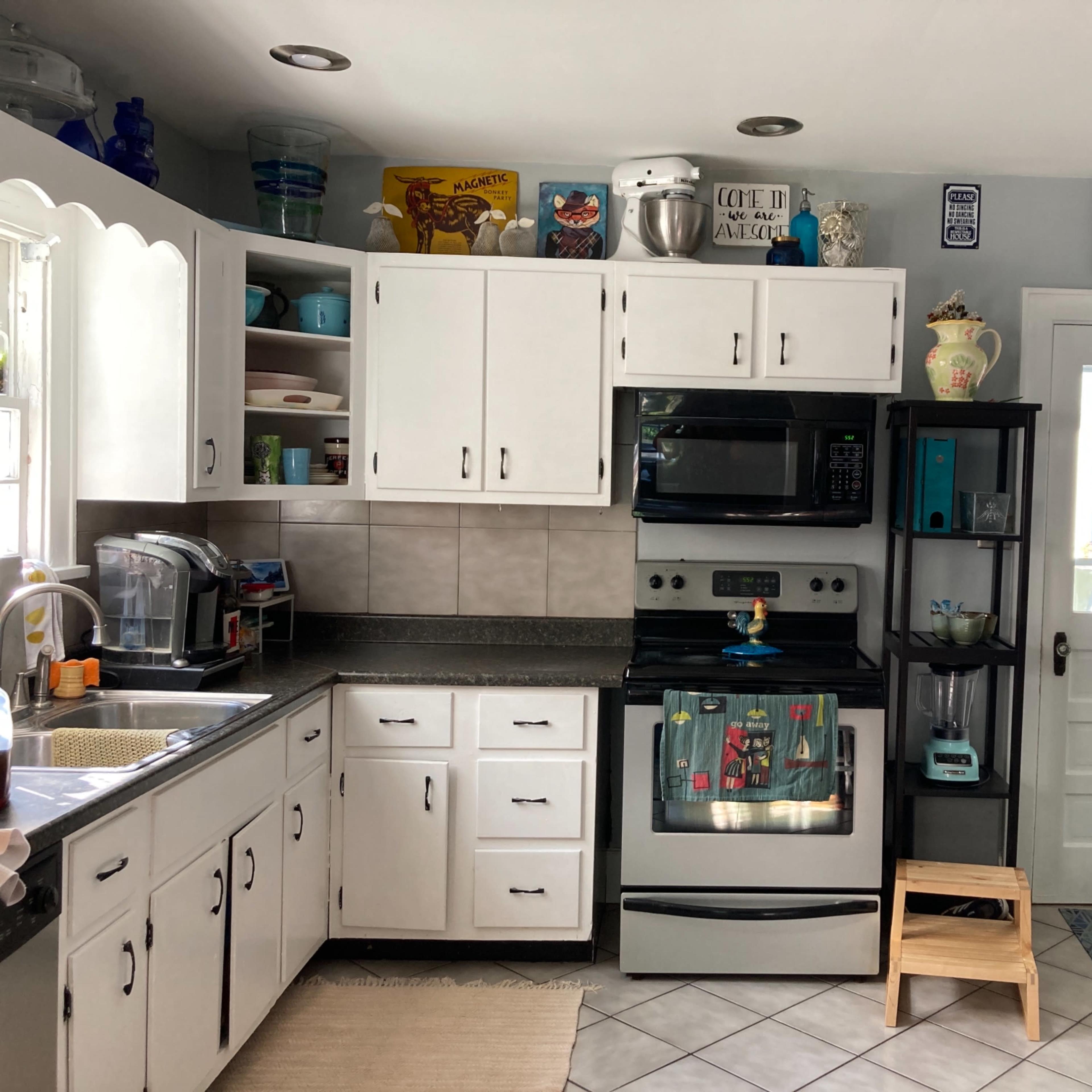 A compact kitchen featuring white cabinets, a black stove and microwave, and various appliances on the counter and shelves.