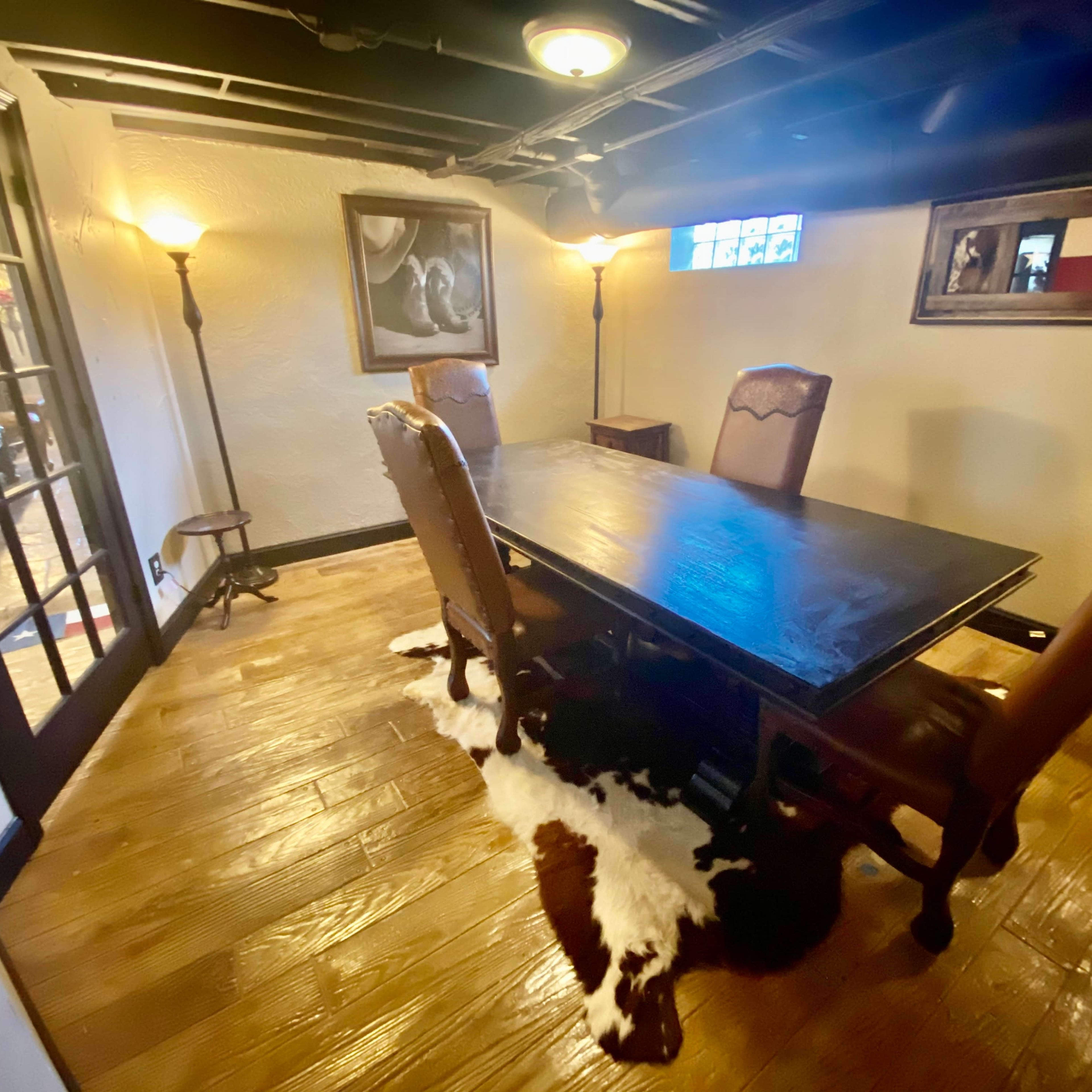 A dimly lit room with a large wooden table surrounded by four leather chairs, a cowhide rug on the floor, and framed artwork on the walls.