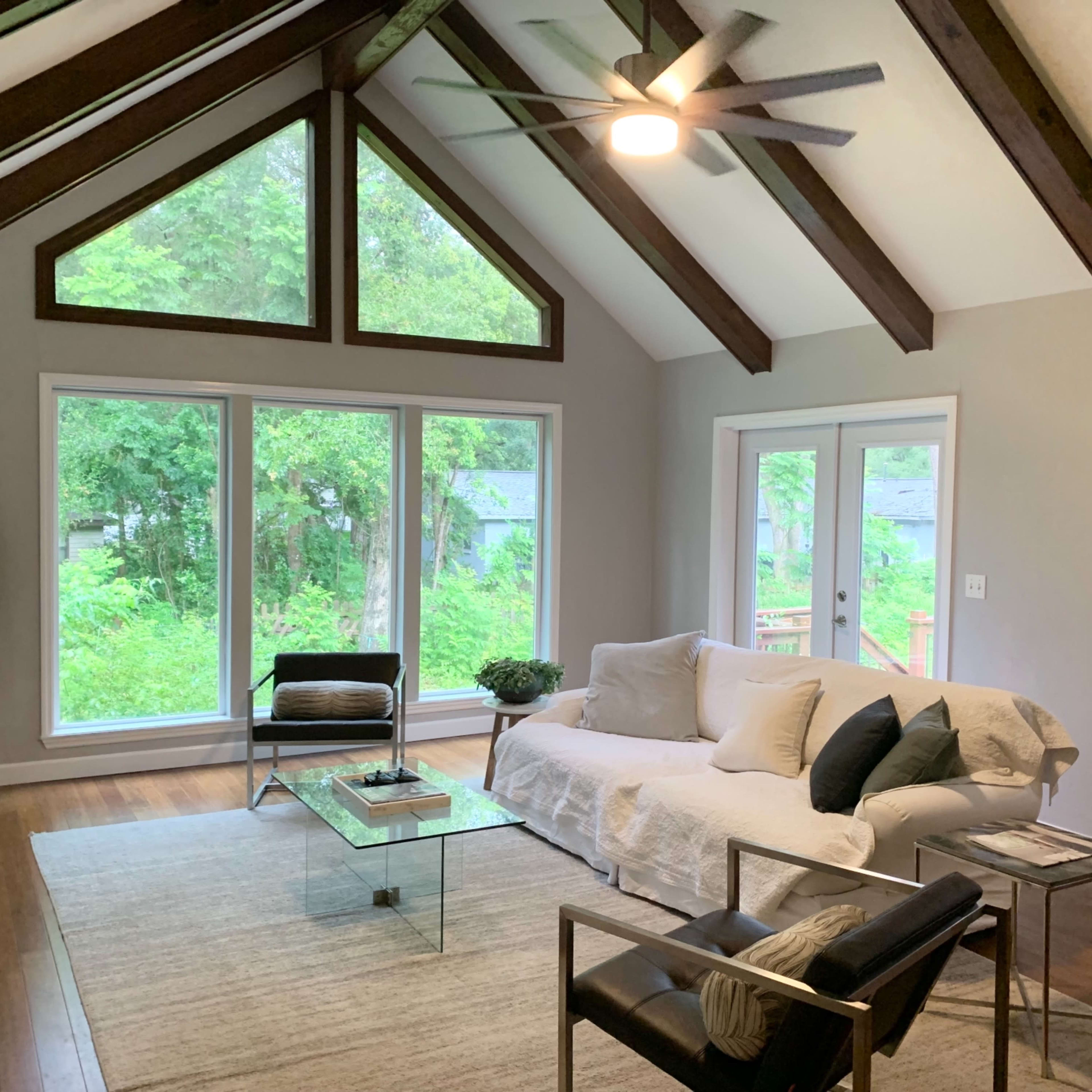 A modern living room featuring a large window with a view of greenery, a light-colored sofa, a glass coffee table, and wooden beams on the ceiling.