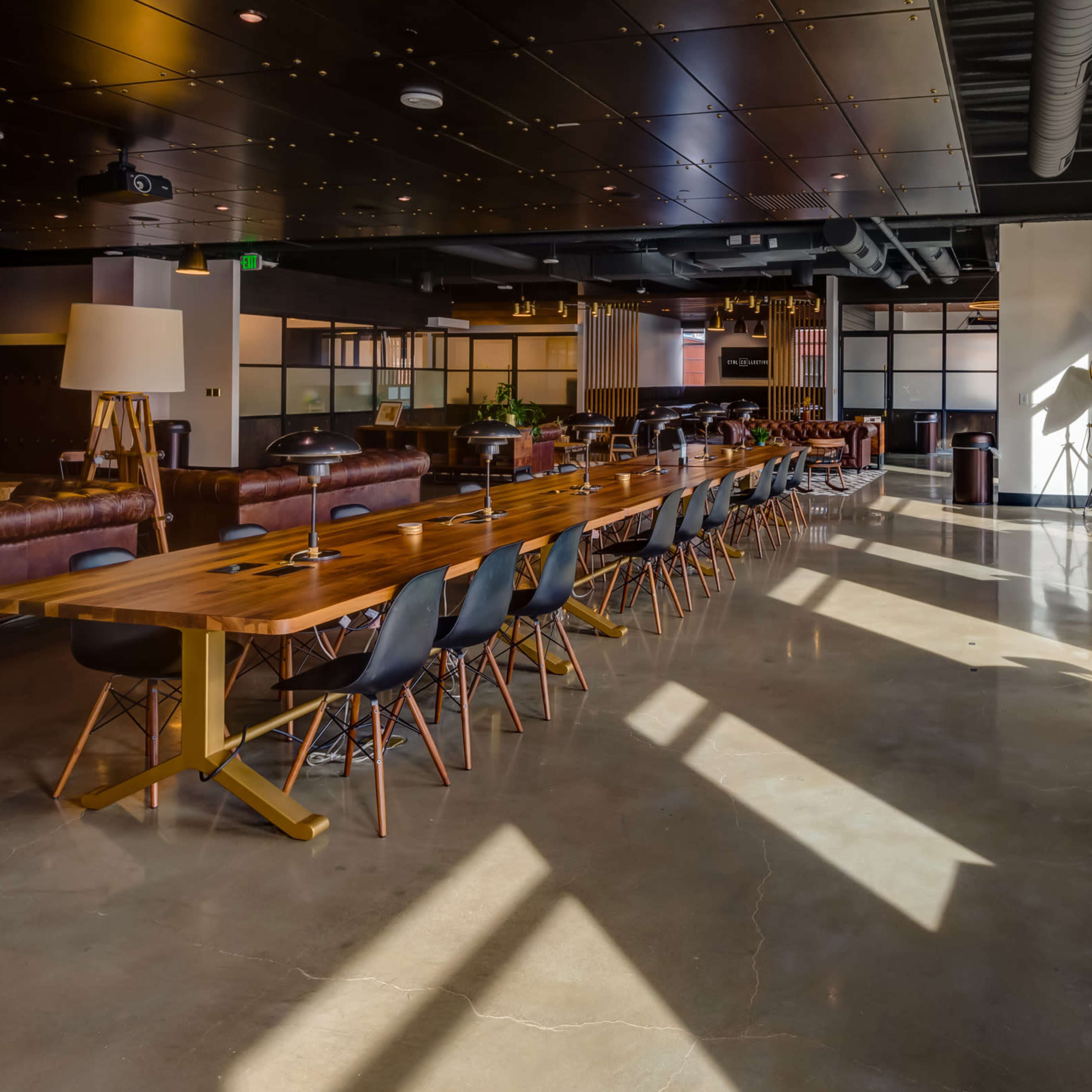 A modern open workspace with a long wooden table, high-backed chairs, and various seating areas bathed in natural light from large windows.