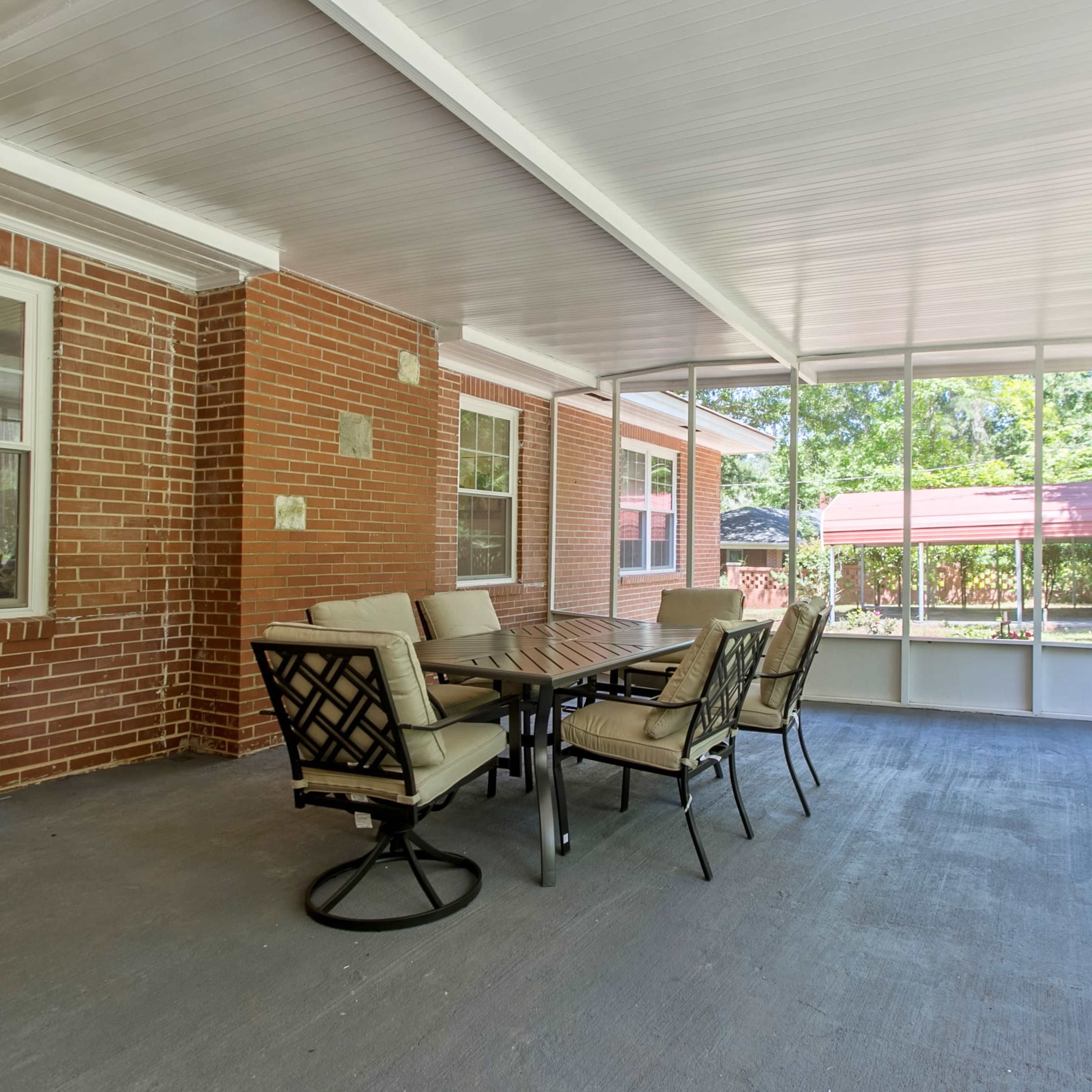 A screened-in porch featuring a rectangular dining table surrounded by six chairs on a concrete floor, with brick walls and large windows allowing views of the outdoors.