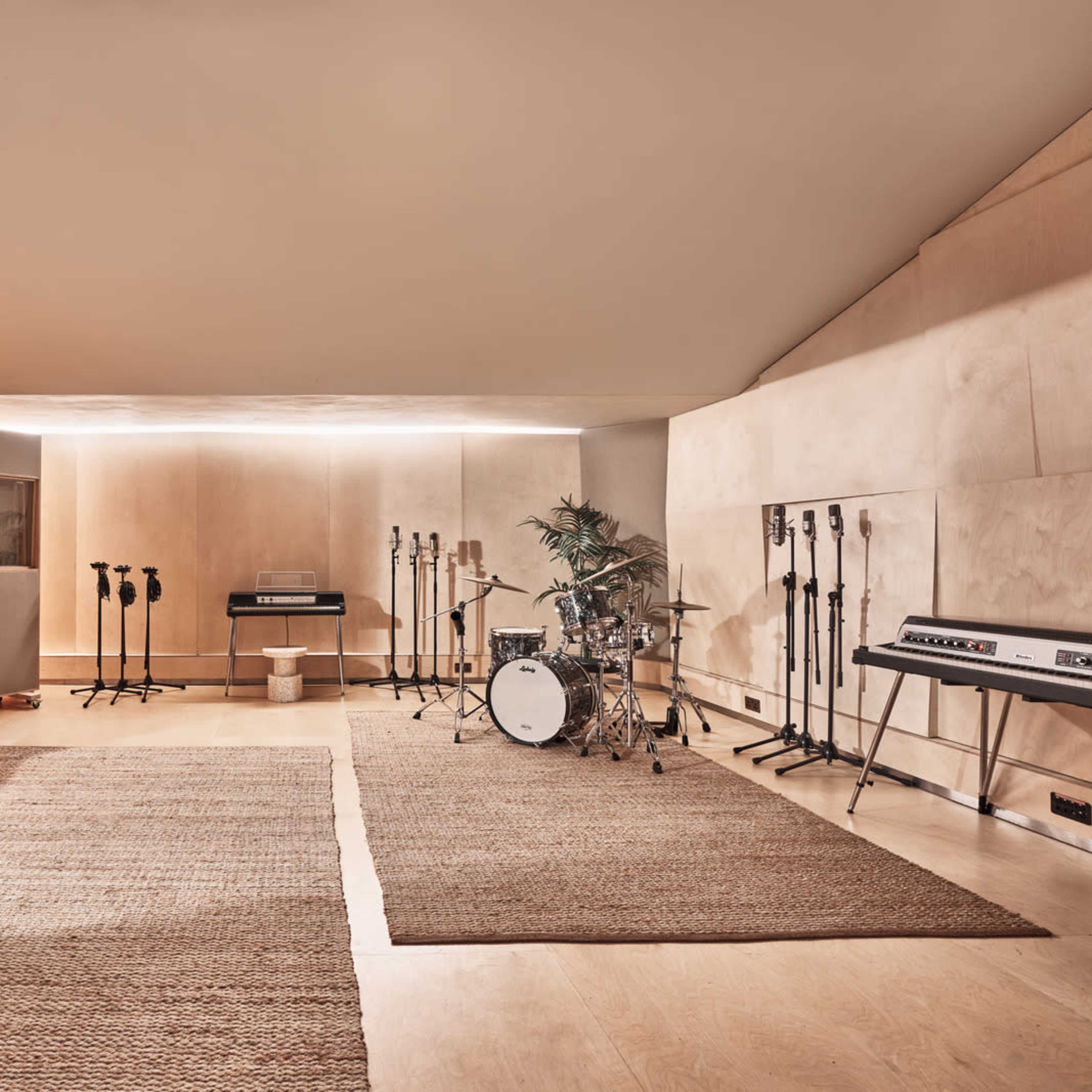 A spacious music studio features a drum set, keyboards, and multiple microphones arranged on stands, with a large window and natural light illuminating the room.