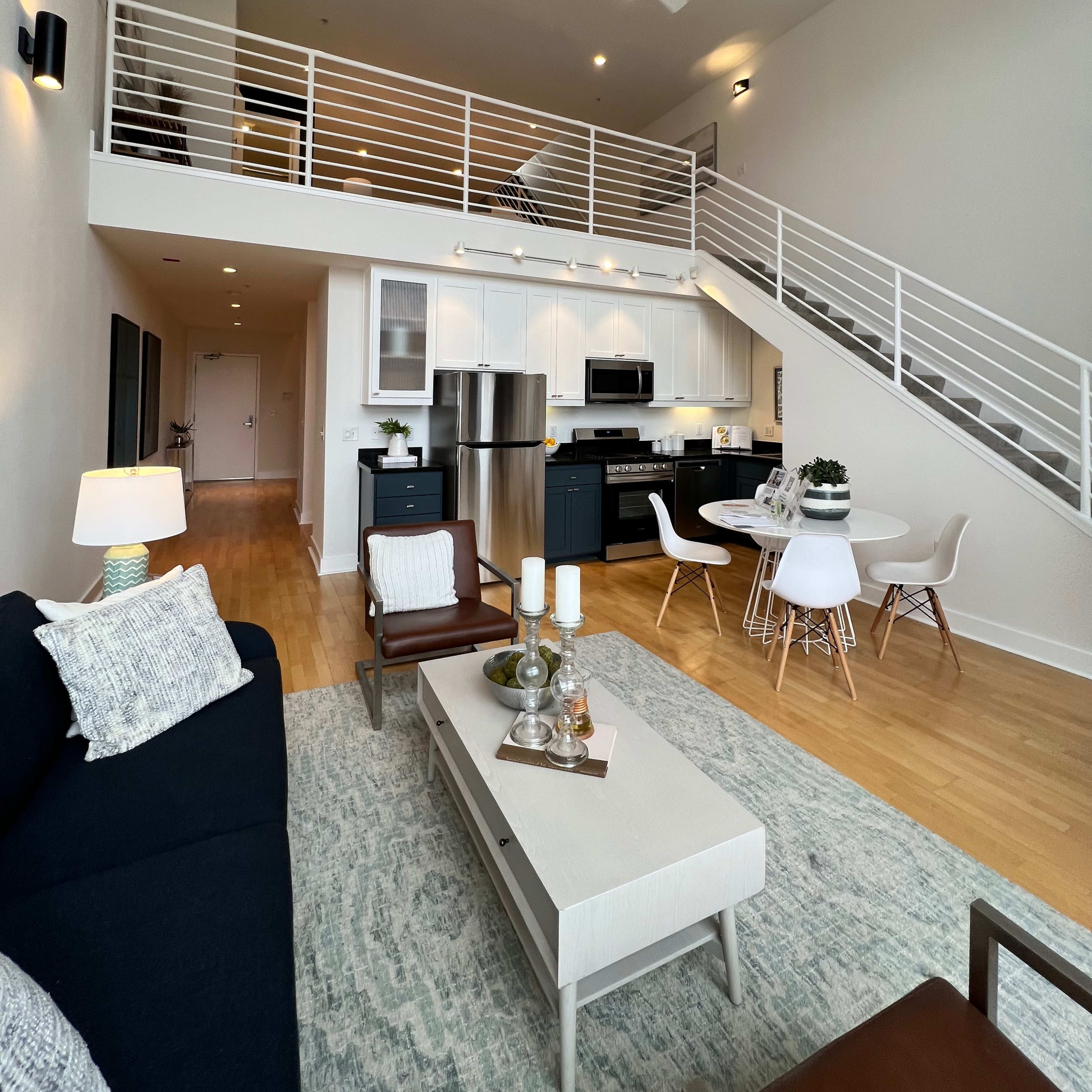 A modern, two-level apartment interior featuring a living area with a sofa, coffee table, and dining space, along with a staircase leading to an upper level.