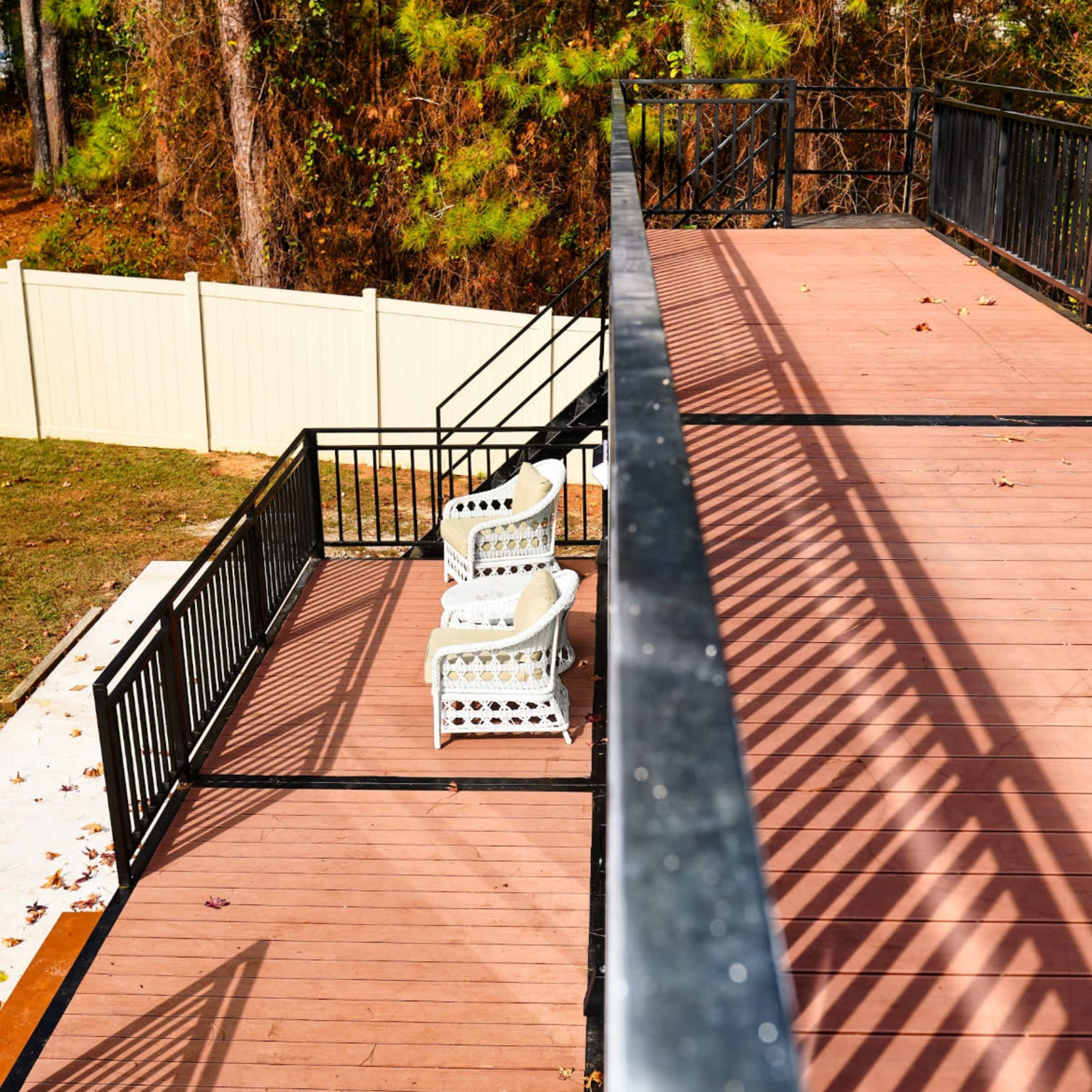A second-story deck features two white chairs surrounded by a wooden surface and a railing with shadows cast by the sunlight.