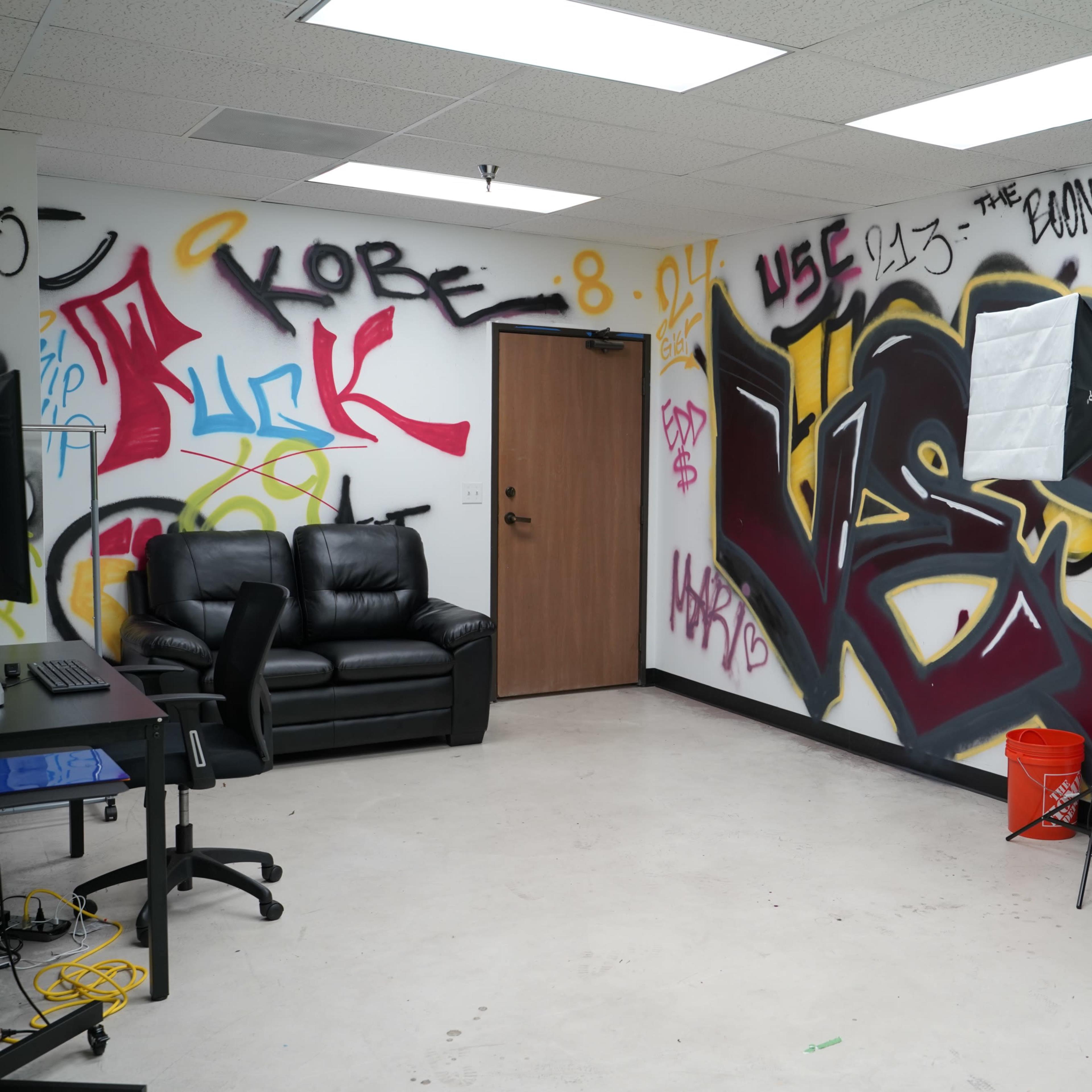 The room features graffiti-covered walls, a black leather couch, and photography lighting equipment set up in a sparse space.
